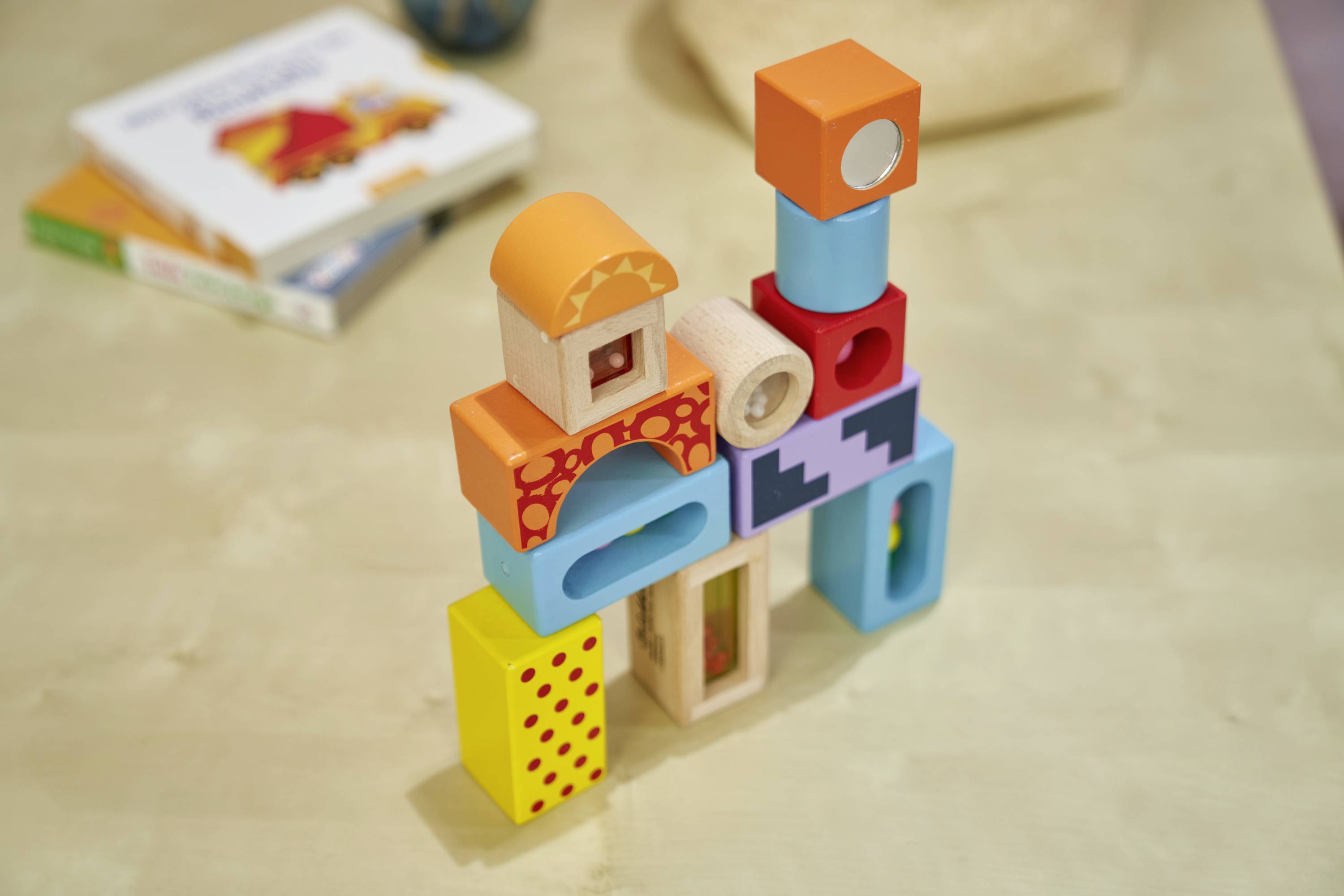 Bunte Holzbausteine sind zu einem Turm gestapelt, im Hintergrund liegen unscharfe Bücher auf einem Tisch.