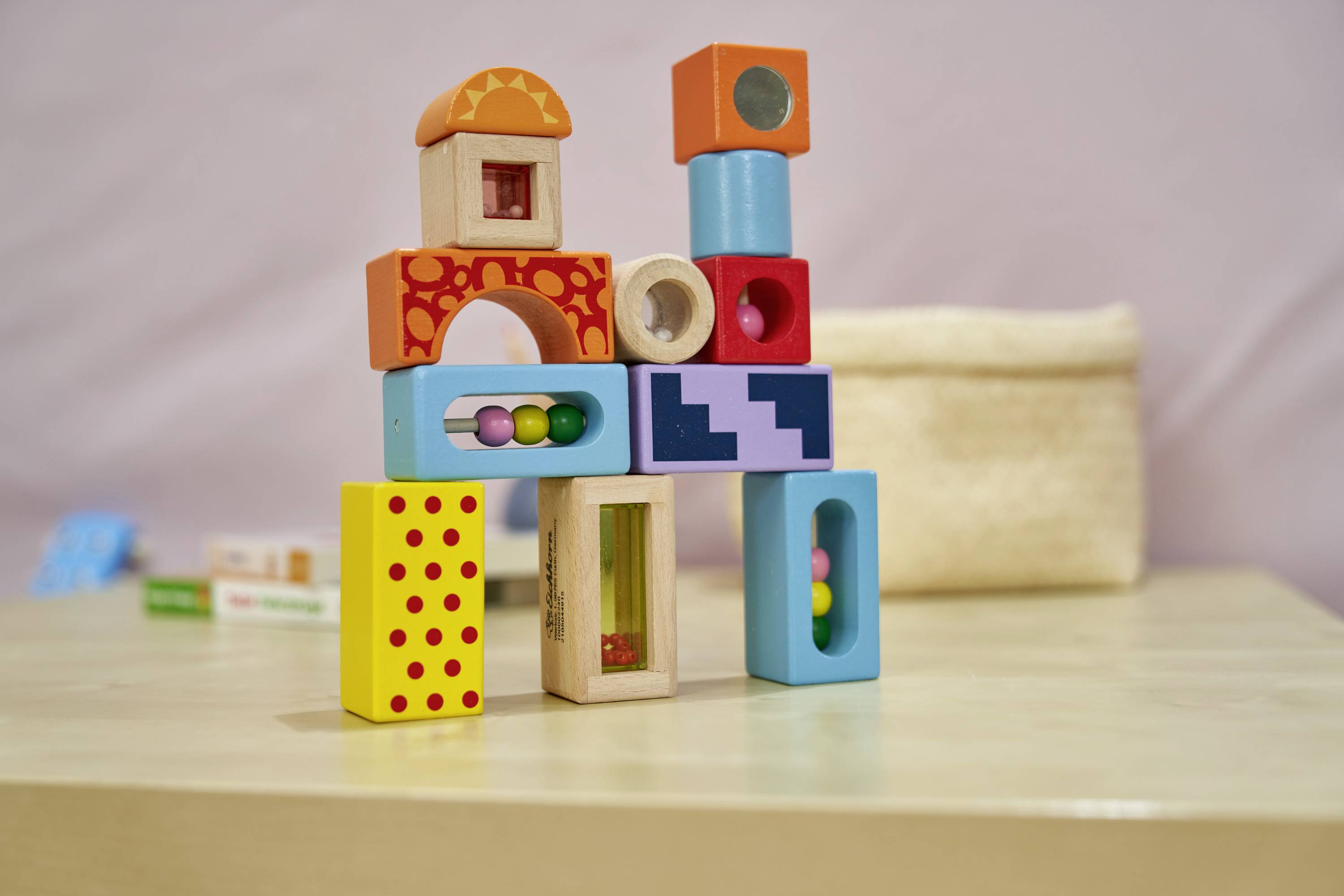 Bunte Holzbauklötze sind kreativ zu einem kleinen Turm gestapelt, im Hintergrund ein verschwommenes Spielzeug im Regal.
