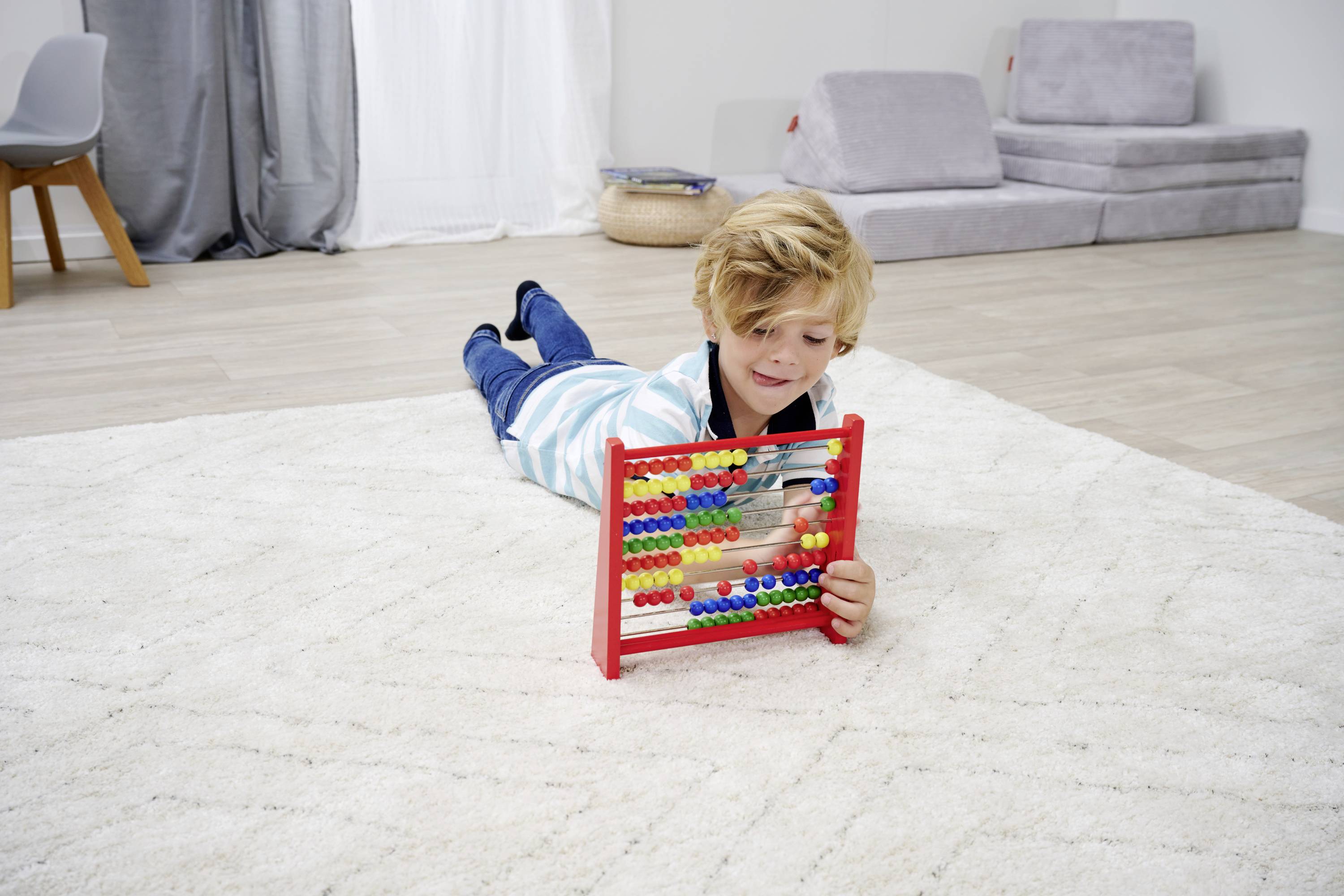Ein Kind liegt auf einem Teppich im Wohnzimmer und spielt mit einem roten Abakus. Im Hintergrund sind ein Stuhl und ein Sofa zu sehen.