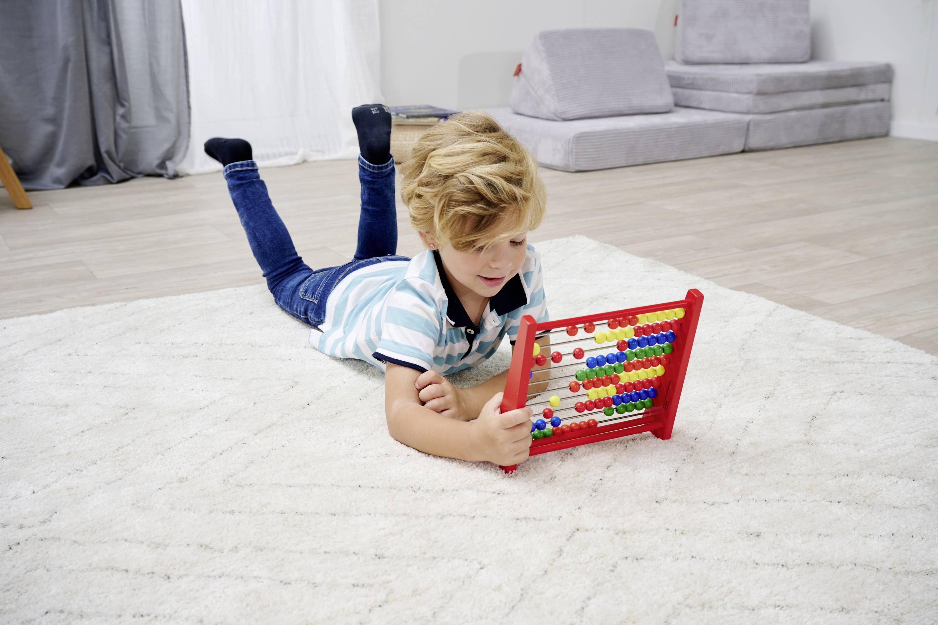 Ein Junge liegt auf einem Teppich und spielt mit einem roten Abakus. Im Hintergrund sind Sofas und Vorhänge zu sehen.