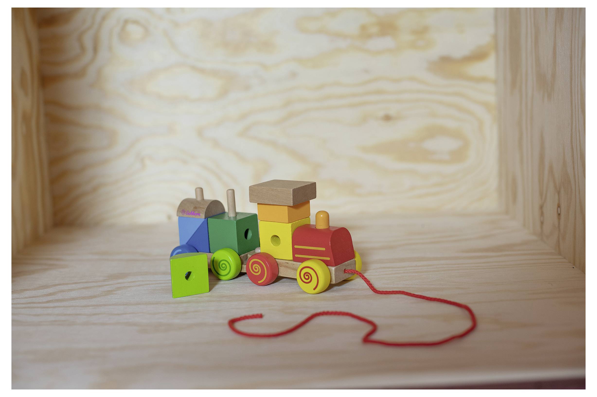 Ein bunter Holzspielzeugzug mit roter Schnur steht auf einem hölzernen Regal. Der Zug hat verschiedene Formen und Farben.