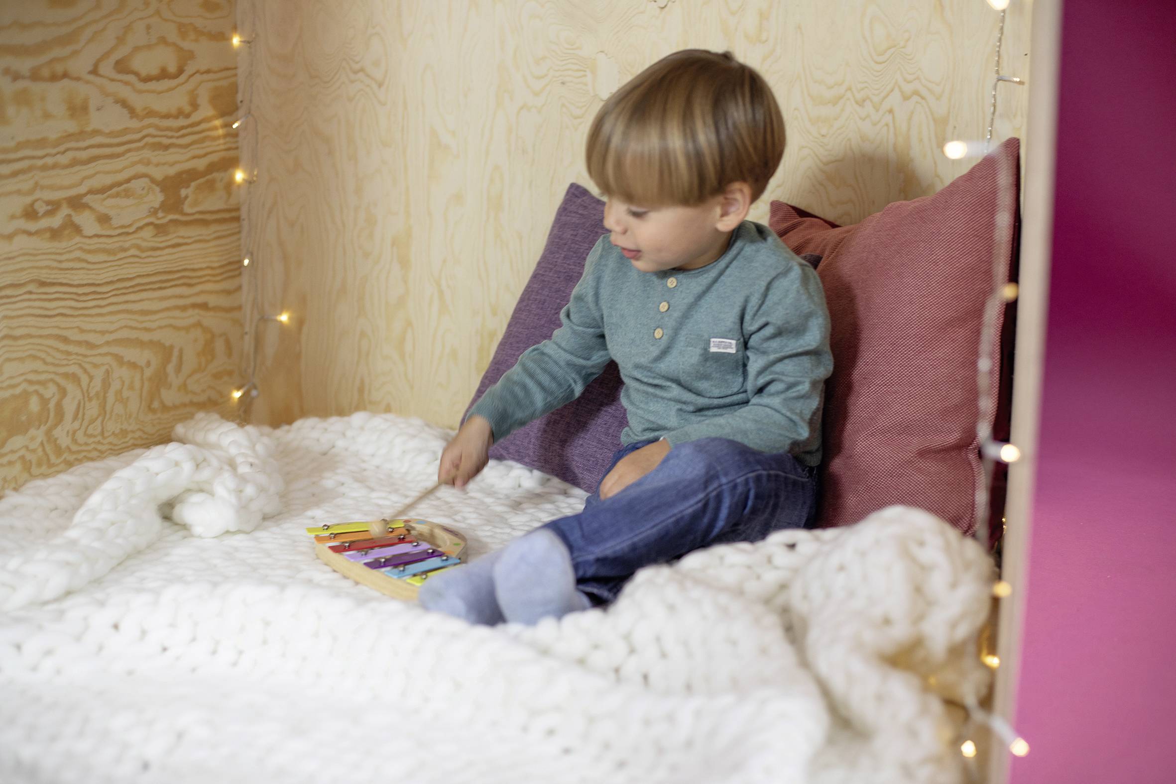 Ein Kind sitzt in einer gemütlichen Ecke mit Kissen und einer Decke, liest ein buntes Buch. Lichterketten schmücken den Hintergrund.