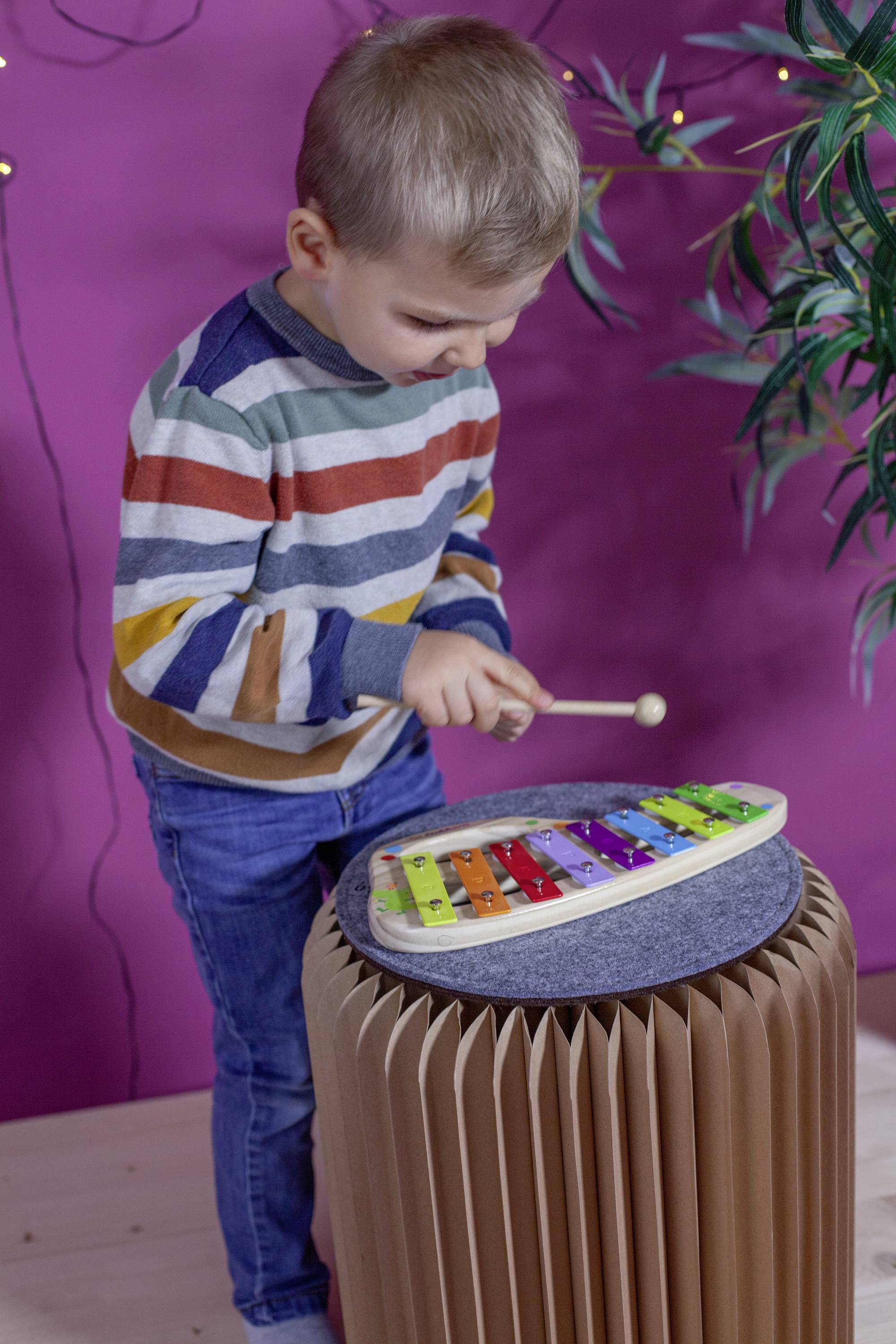 Ein Kind spielt konzentriert ein buntes Xylophon auf einem Papierhocker vor einem lilafarbenen Hintergrund mit Pflanzen.