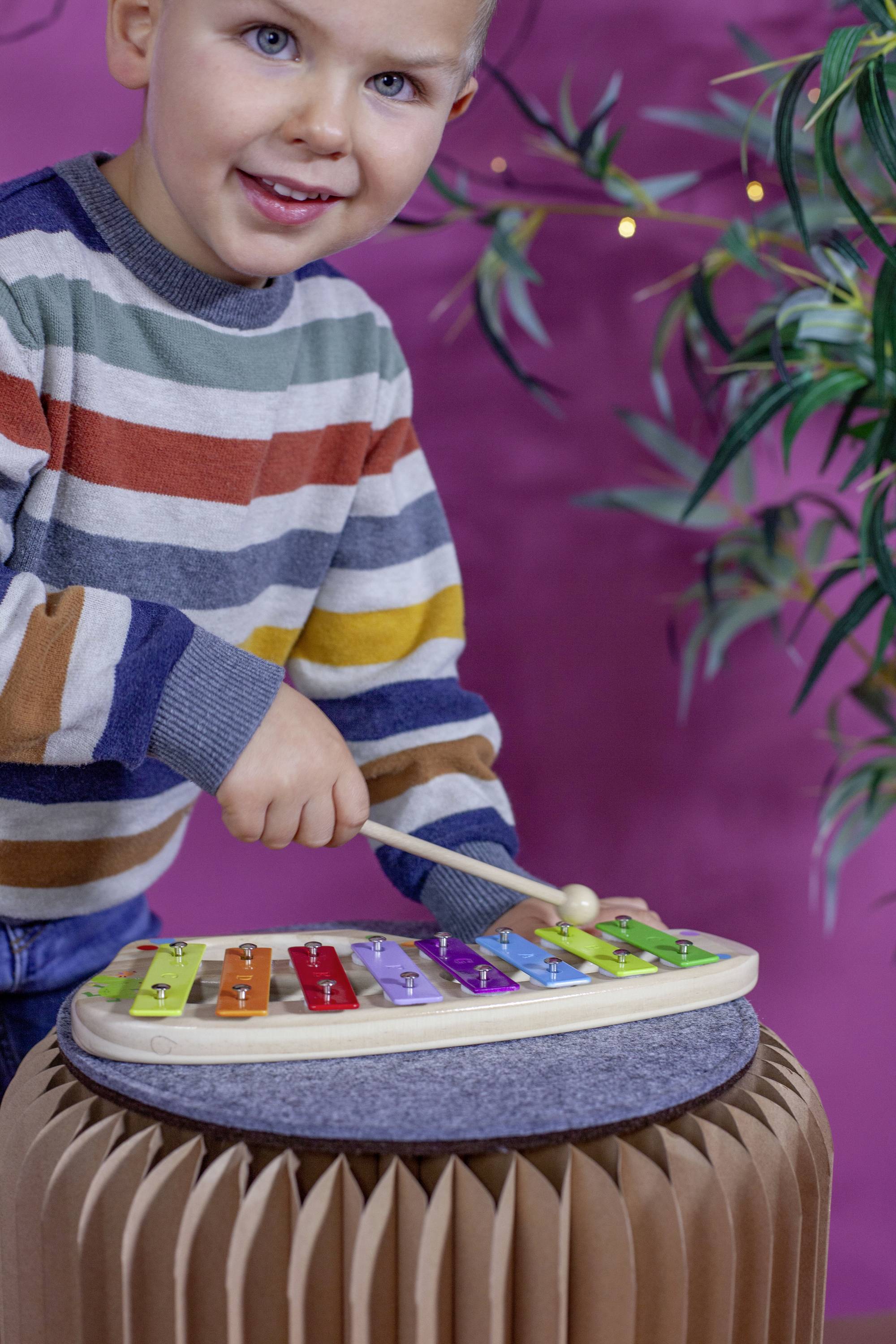 Ein Kind spielt fröhlich ein buntes Xylophon. Im Hintergrund sind Pflanzen und eine rosa Wand zu sehen.