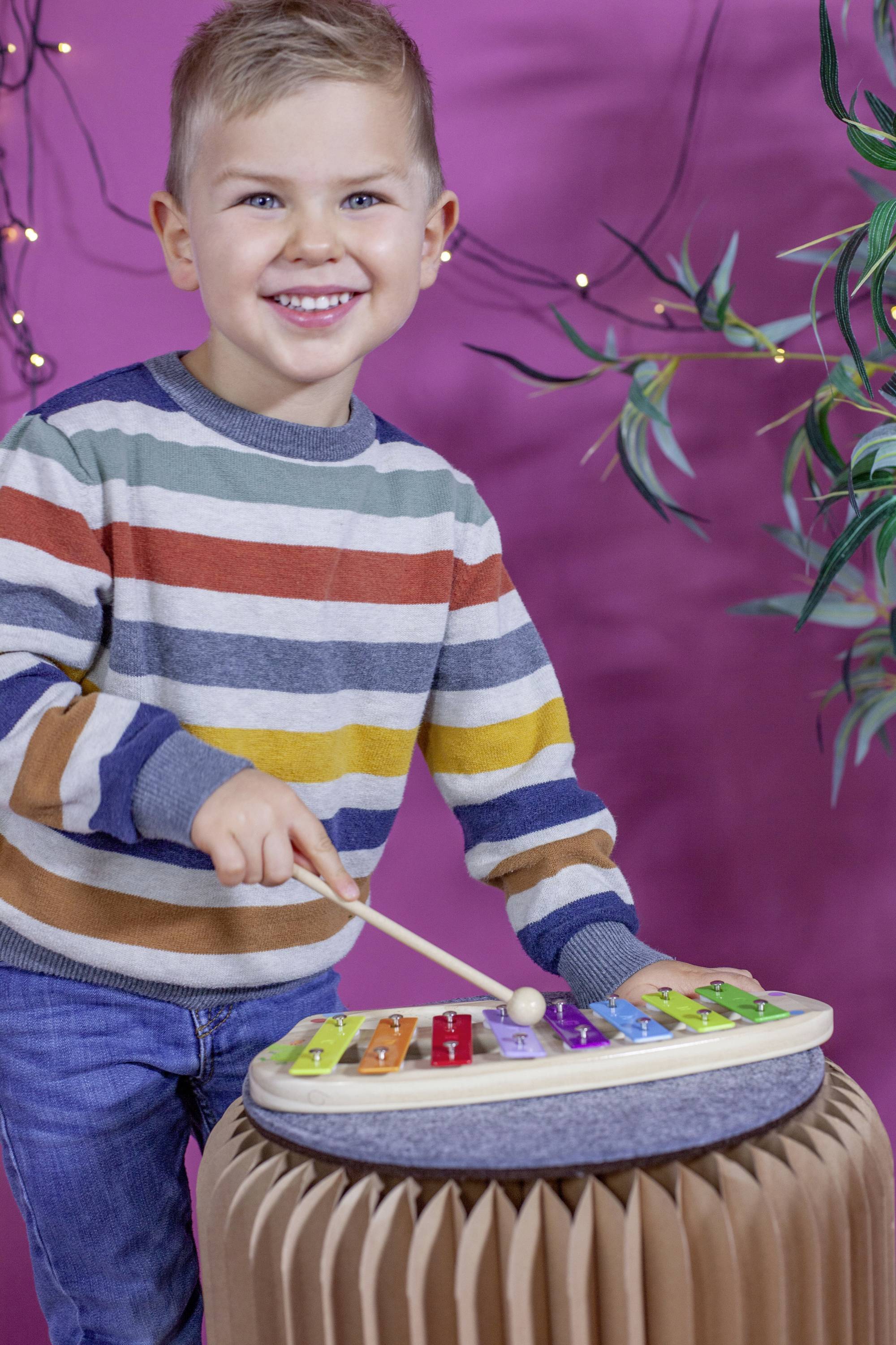 Ein lächelndes Kind spielt ein farbenfrohes Xylophon mit einem Schlägel. Im Hintergrund sind lila und pflanzliche Dekorationen zu sehen.