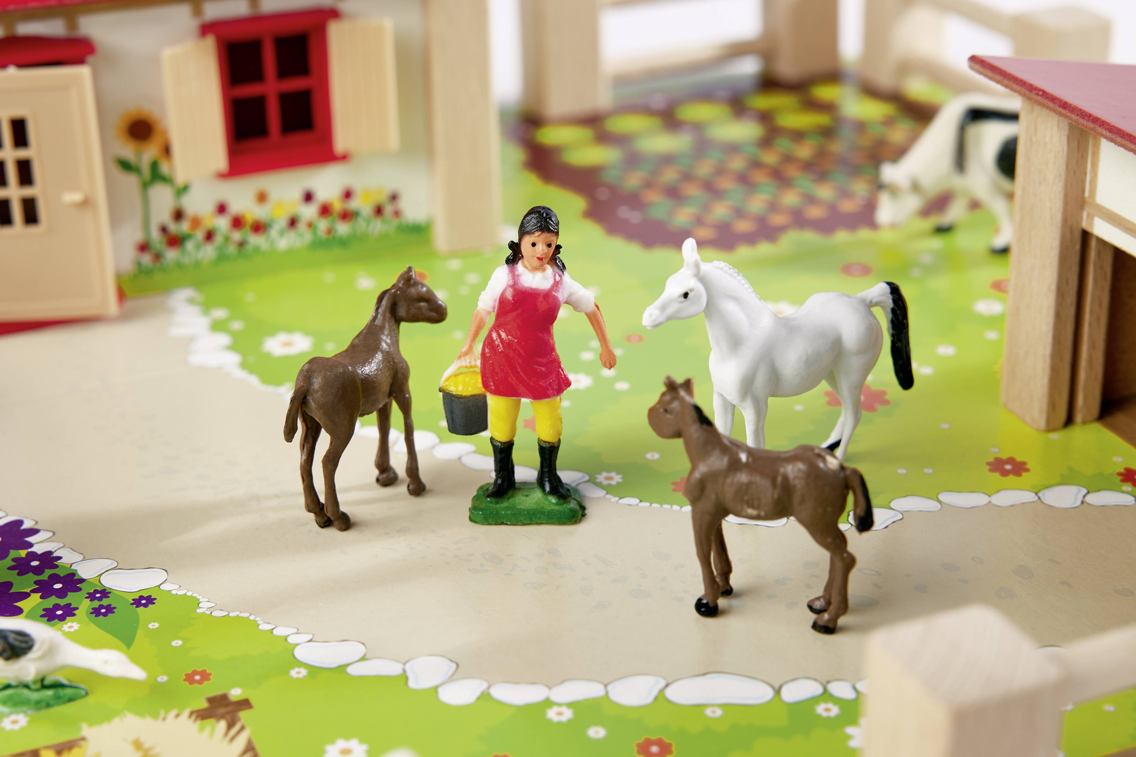 Miniaturbauernhof mit Plastikfiguren: Eine Bäuerin mit Eimer steht in einem Garten, umgeben von drei Pferden. Gebäude und bunte Blumen im Hintergrund.
