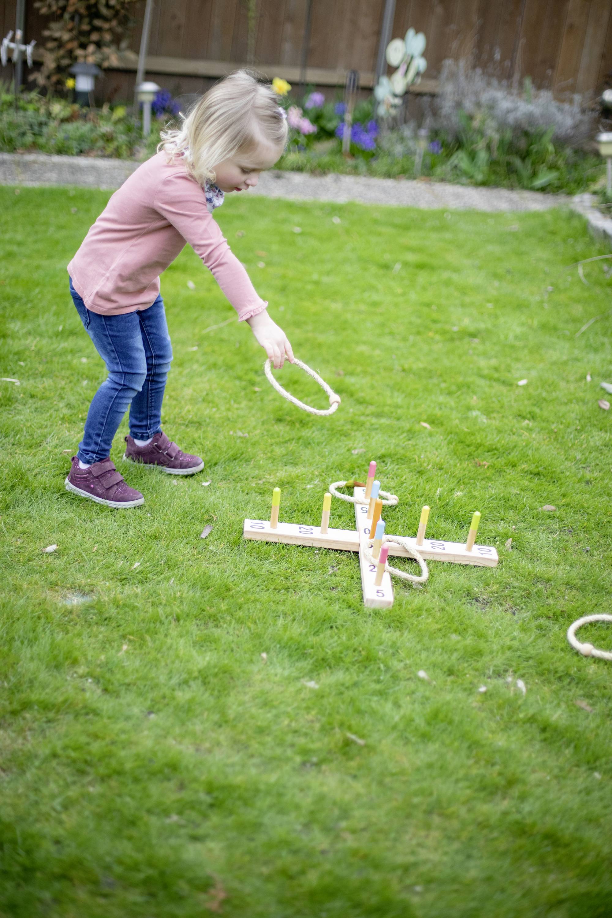 Ein Kind wirft einen Ring bei einem Gartenspiel, versucht, die Stange zu treffen. Im Hintergrund befinden sich Blumenbeete.