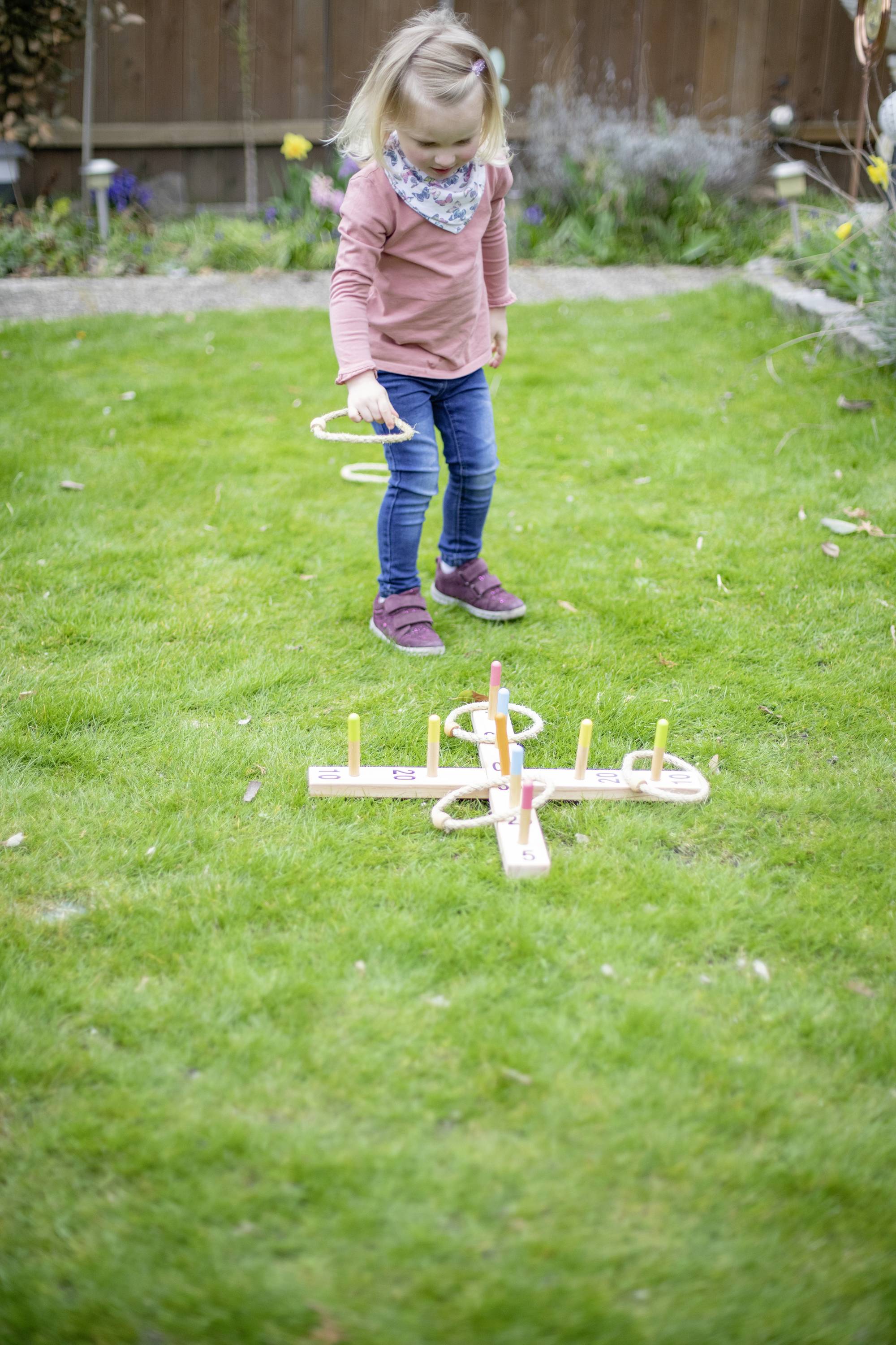 Ein Kind spielt im Garten Ringwurf mit einem Holzspielzeug. Es wirft Ringe auf Stäbe, die auf dem Rasen aufgestellt sind.