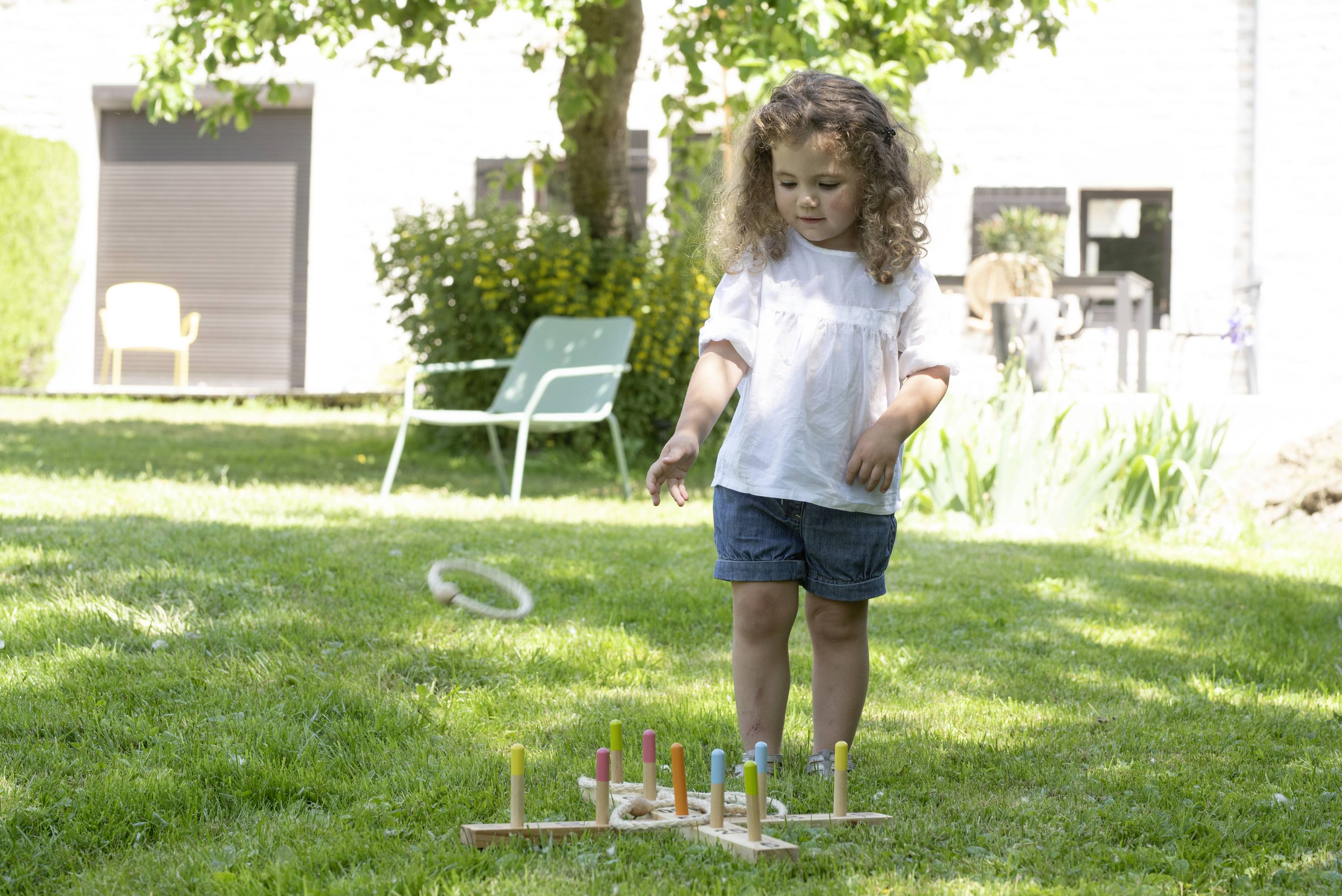 Ein kleines Mädchen spielt im Garten ein Ringwurfspiel. Sie steht auf einer Wiese und zielt auf bunte Stäbe mit Ringen.