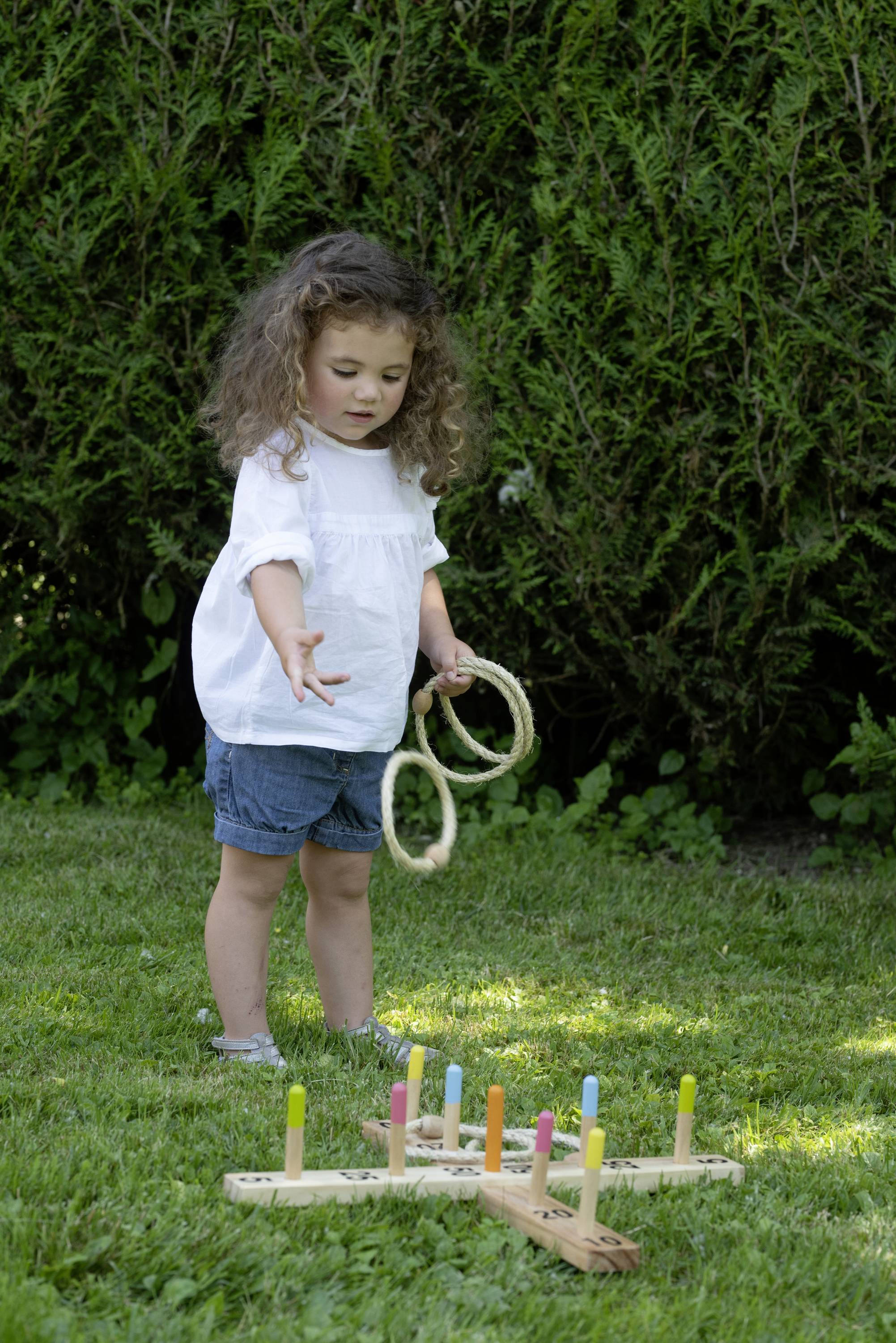 Kleines Mädchen wirft Ringe auf ein Steckspiel im Garten. Sie konzentriert sich auf das Spiel, um die Ringe über die bunten Stäbe zu werfen.