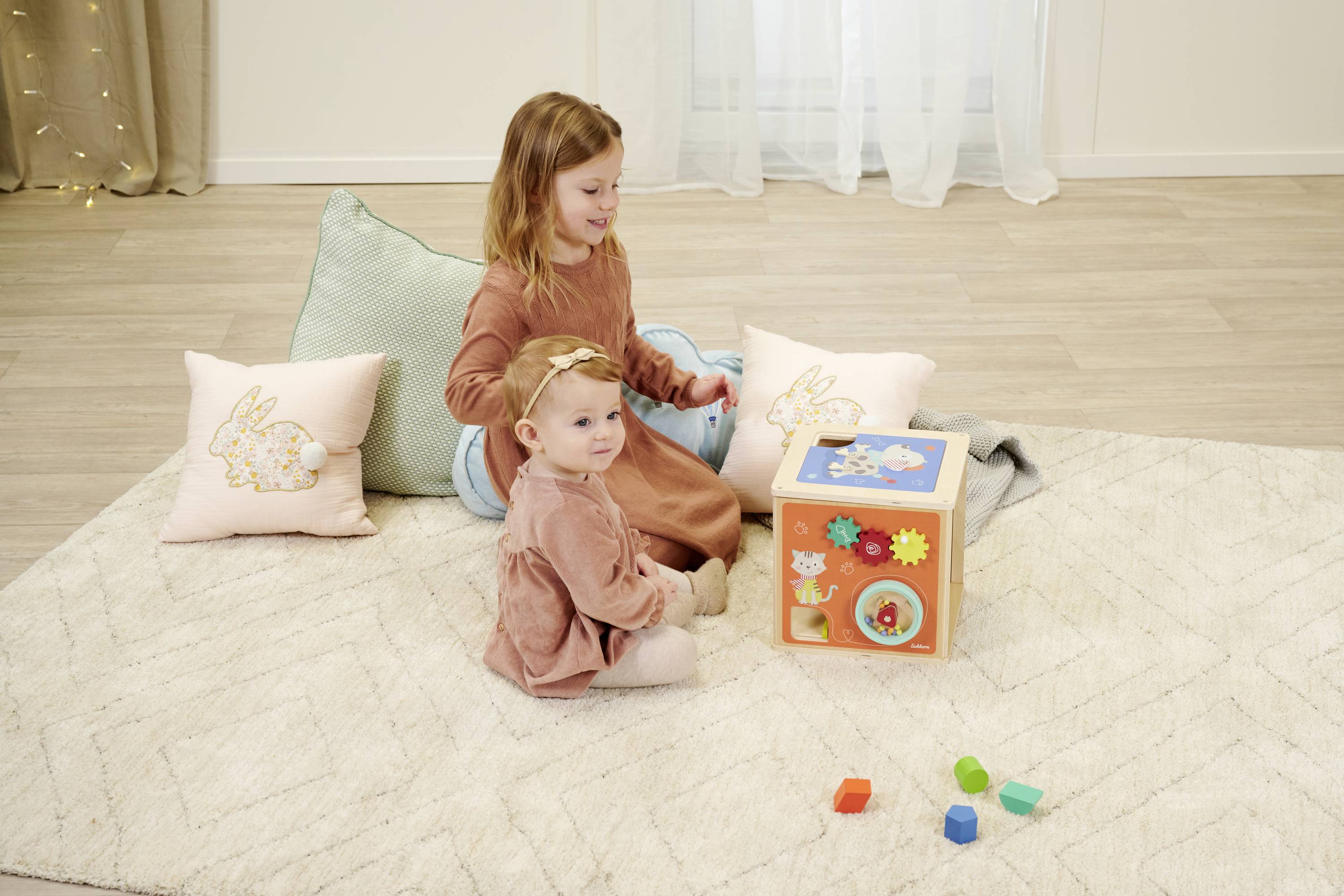 Zwei Mädchen spielen auf einem Teppich mit einem Lernspielzeug. Kissen liegen im Hintergrund. Das Spielzeug hat bunte Formen und Zahnräder.