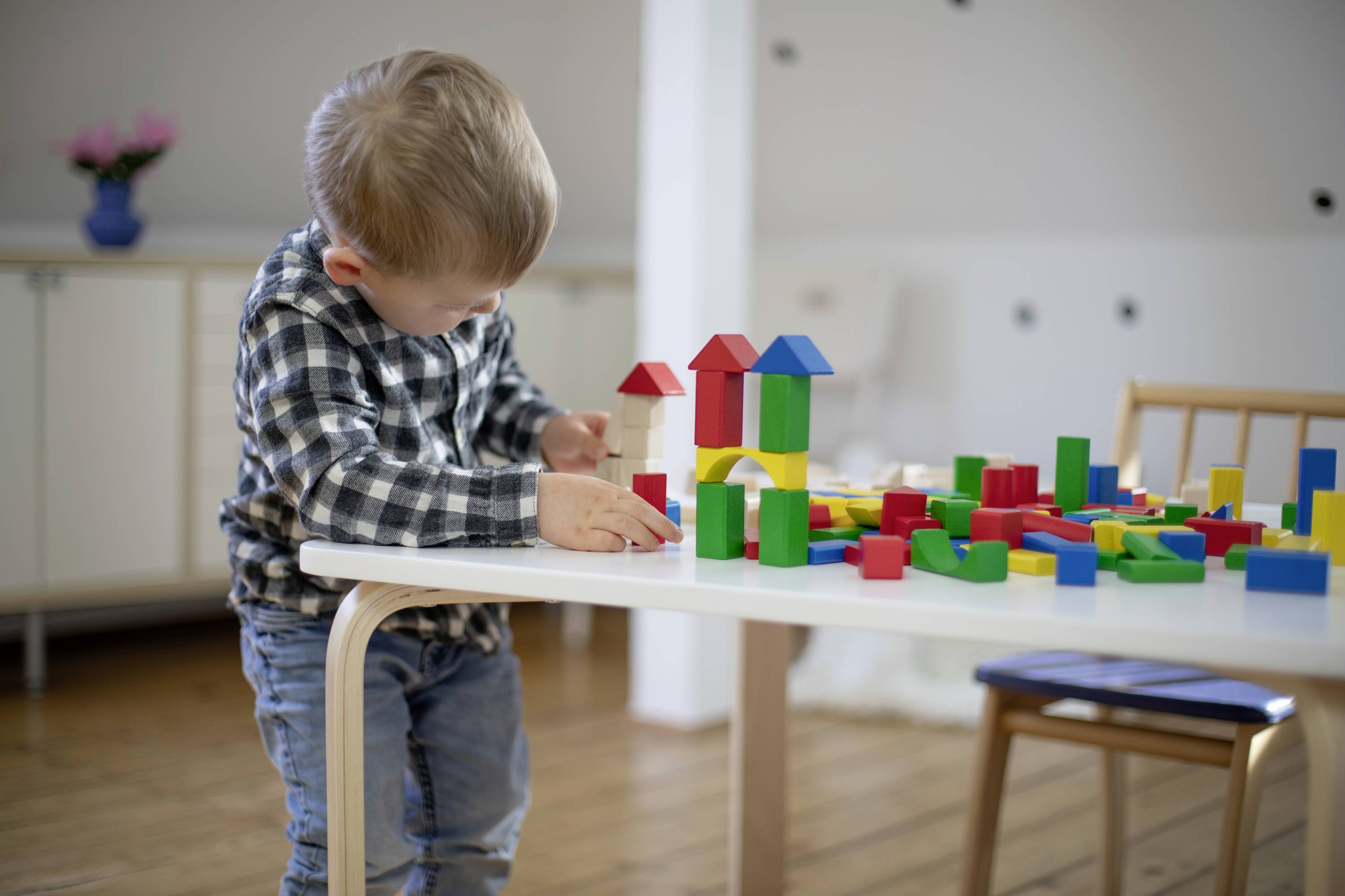 Ein kleiner Junge baut mit bunten Holzklötzen am Tisch. Im Hintergrund sind Möbel zu sehen.