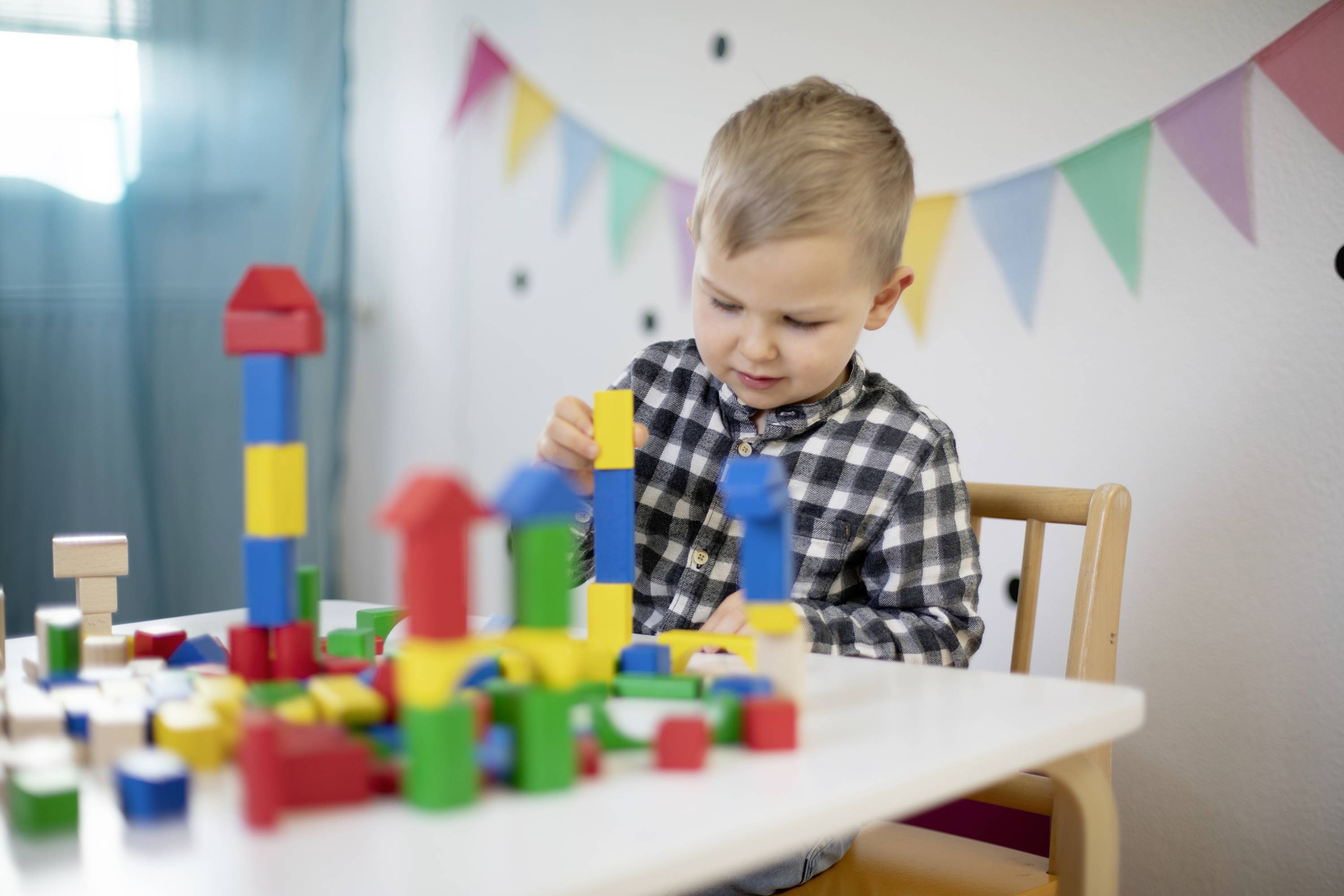 Ein kleines Kind sitzt an einem Tisch und spielt mit bunten Bauklötzen. Im Hintergrund sind bunte Wimpel an der Wand.
