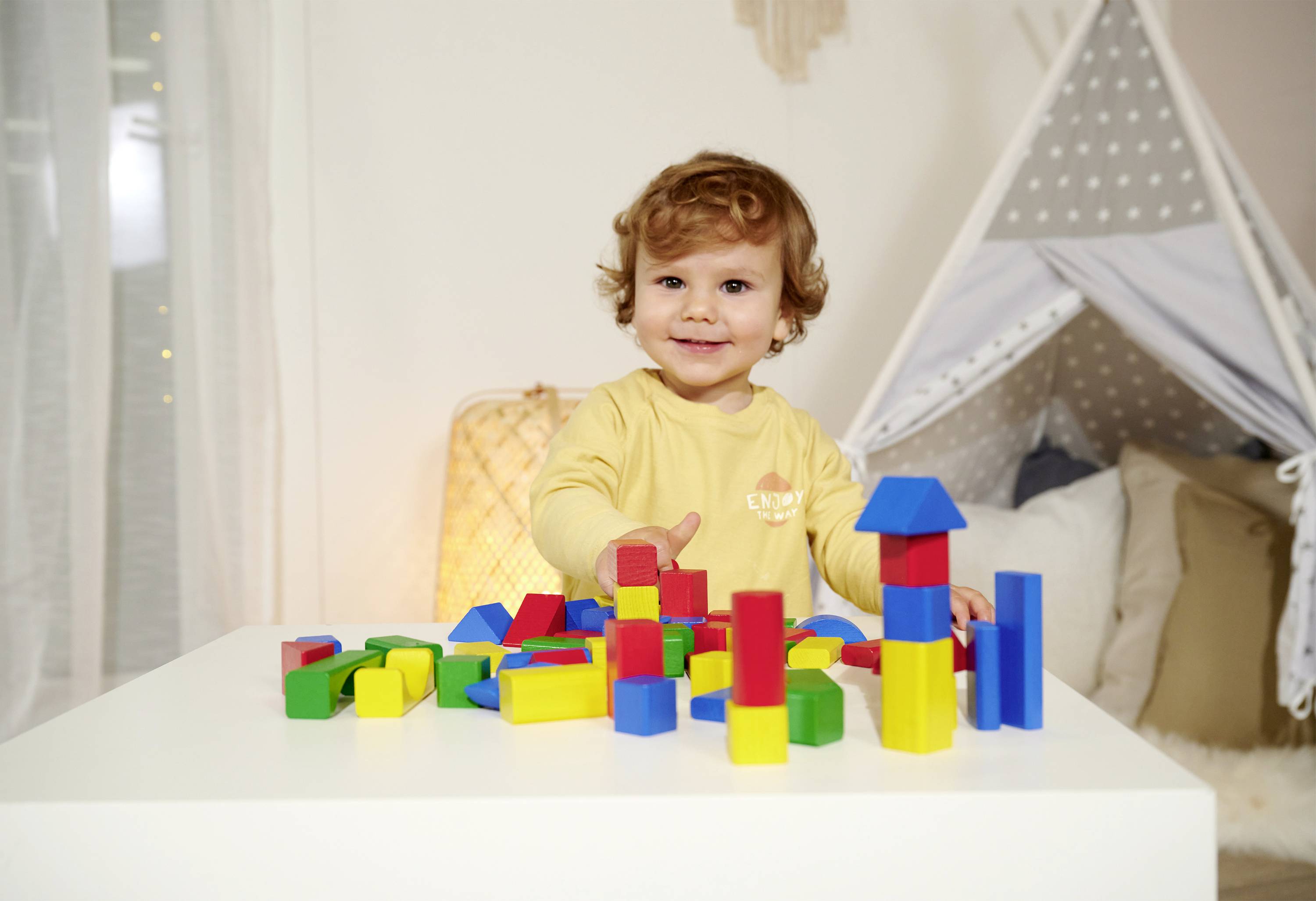 Ein kleines Kind spielt mit bunten Bauklötzen auf einem weißen Tisch vor einem Spielzelt im Hintergrund.
