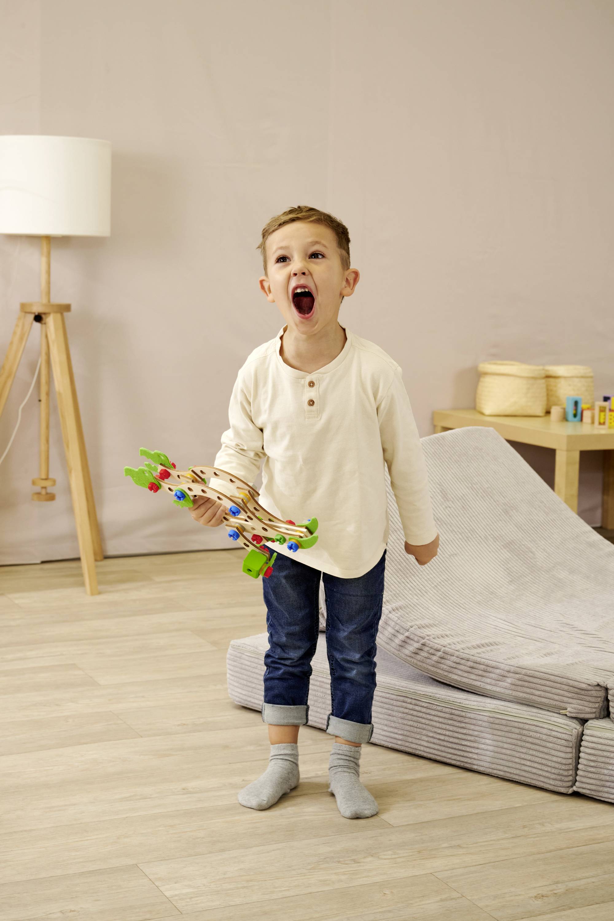Ein Kind in einem Raum ruft freudig und hält ein buntes Spielzeugflugzeug. Im Hintergrund sind eine Lampe und ein Stuhl zu sehen.