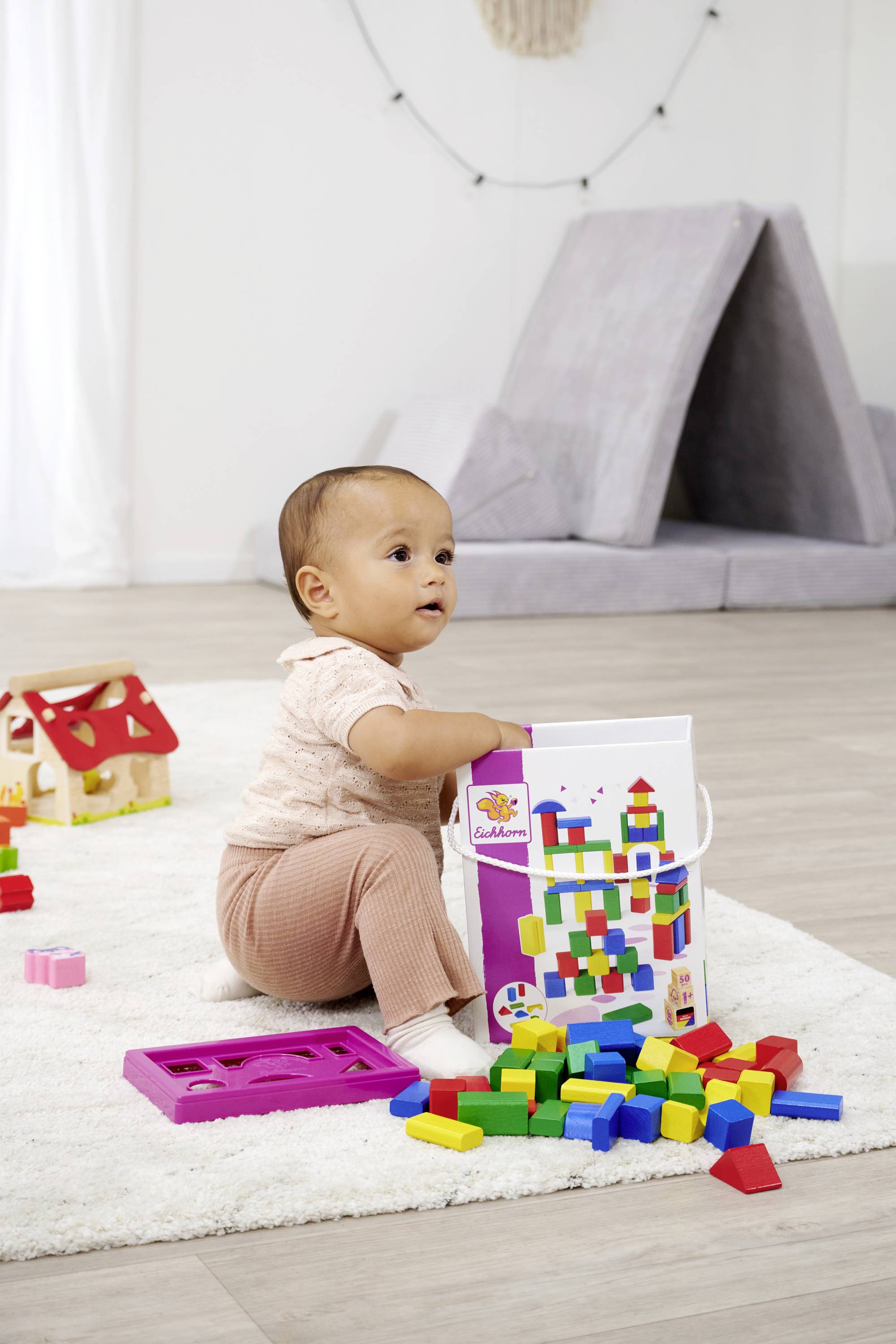 Ein Kleinkind spielt auf einem Teppich mit bunten Bauklötzen, die aus einem Eimer stammen. Im Hintergrund steht ein kleines Zelt.