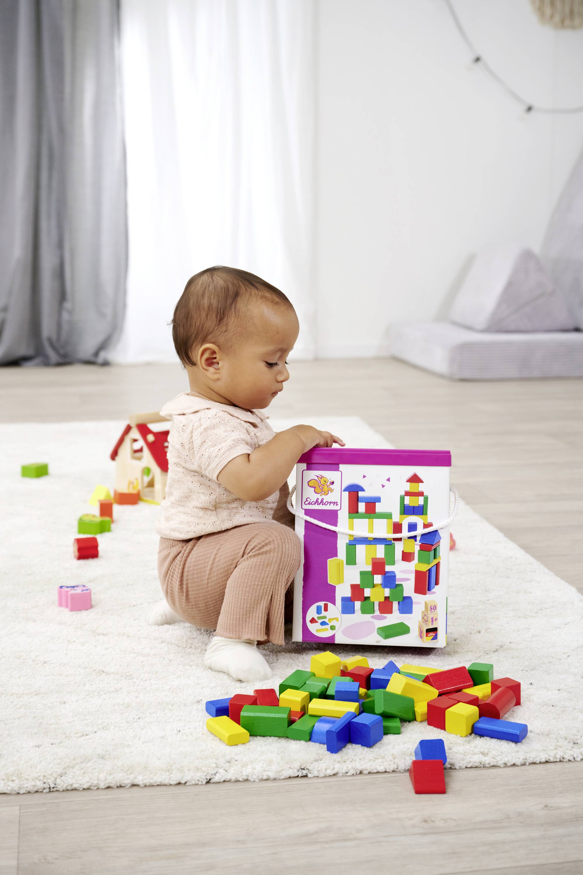 Ein Kleinkind spielt auf einem Teppichboden mit bunten Bauklötzen, während es interessiert eine Schachtel betrachtet.