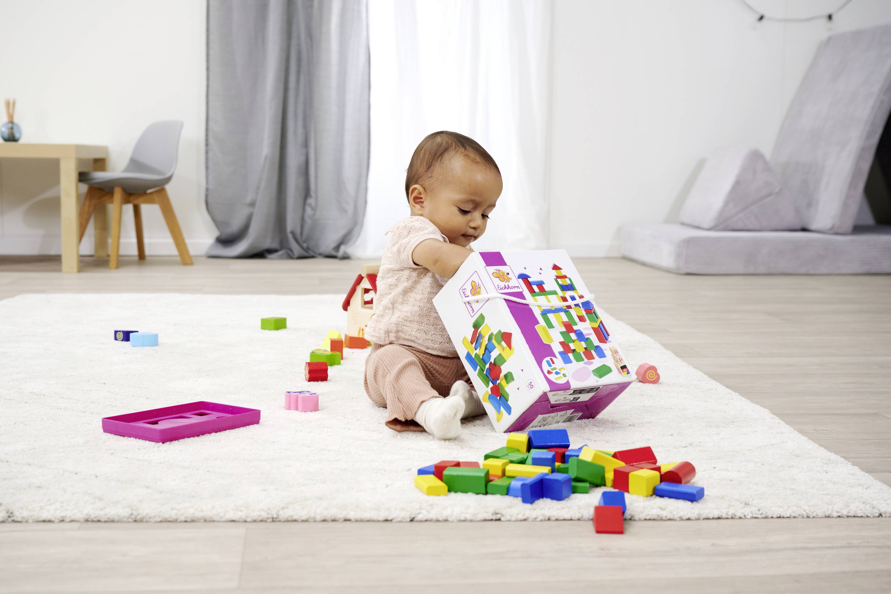 Ein Kleinkind spielt auf einem Teppich mit bunten Bauklötzen und hält eine Schachtel. Im Hintergrund sind Möbel sichtbar.
