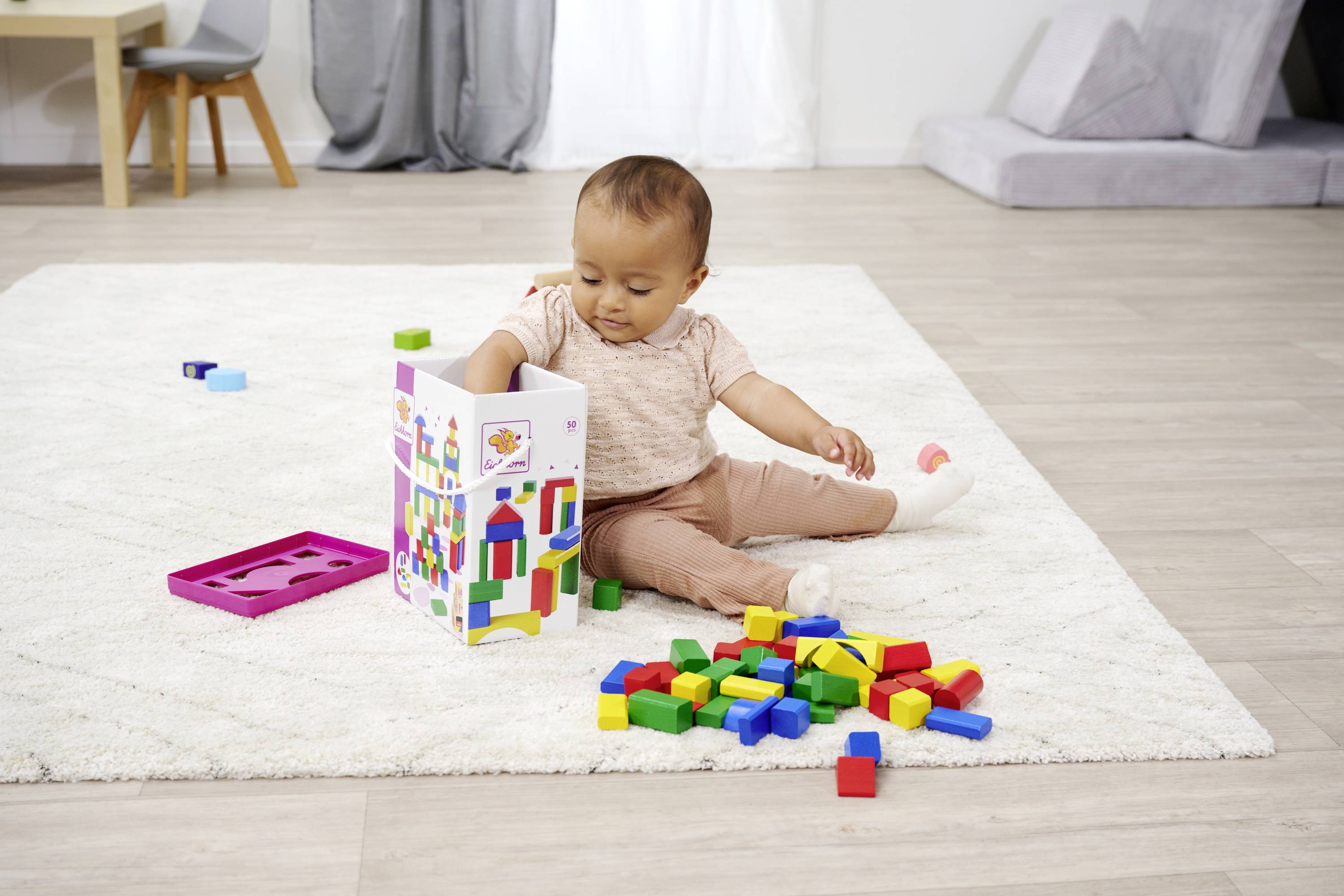 Ein Kleinkind sitzt auf einem Teppich und spielt mit bunten Bauklötzen, die aus einer Box ausgeschüttet wurden.