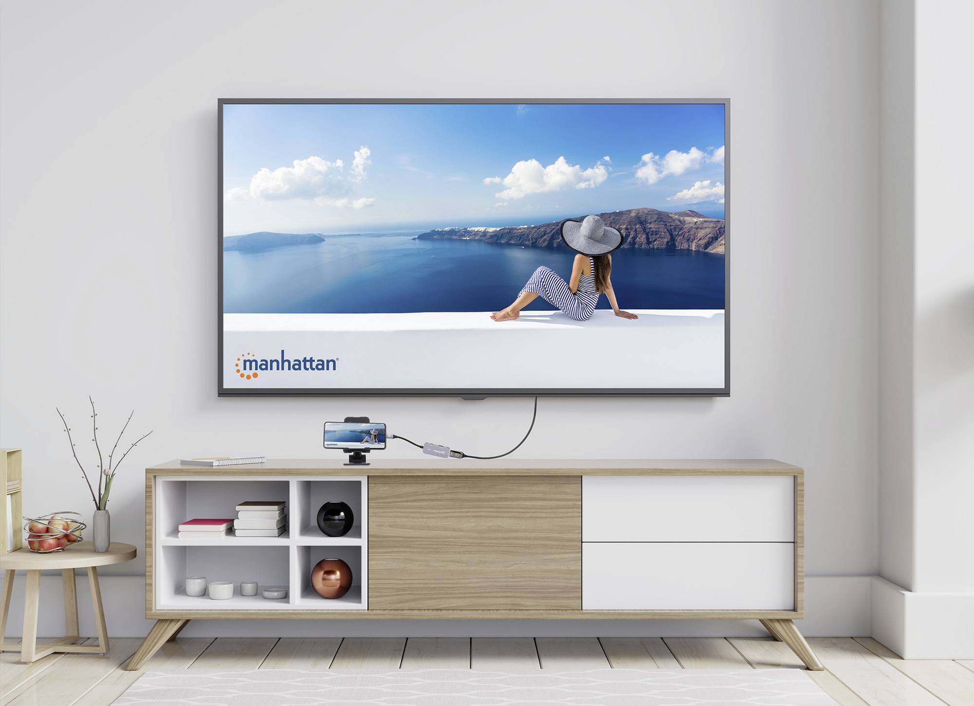 Ein Wohnzimmer mit modernem Fernseher an der Wand. Der Bildschirm zeigt eine Frau mit Hut am Meer. Darunter ein Sideboard mit Deko.
