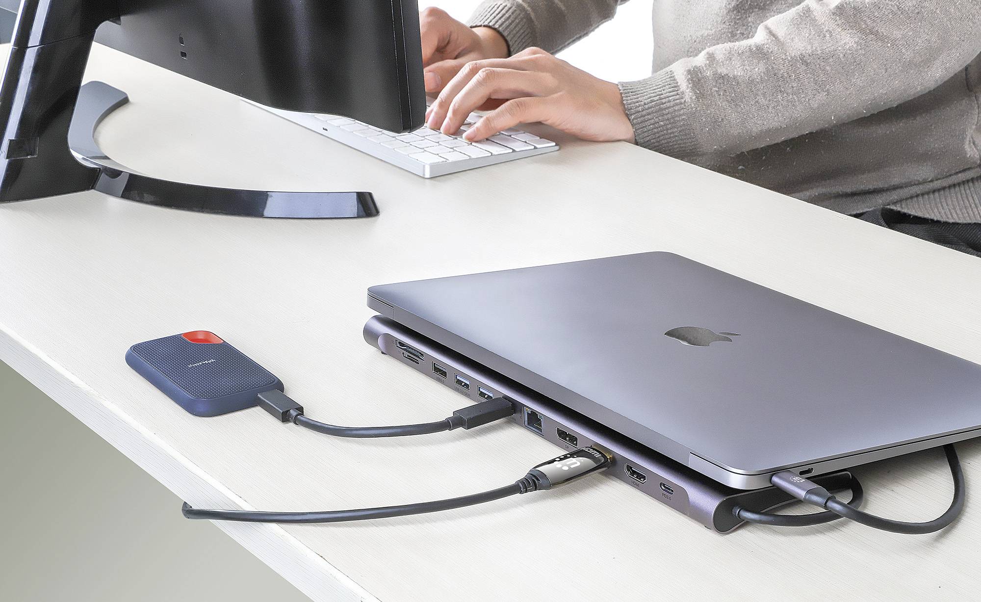 Ein Laptop auf einem Schreibtisch ist mit einem externen Speichergerät und einem Monitor verbunden. Eine Person tippt auf einer Tastatur.