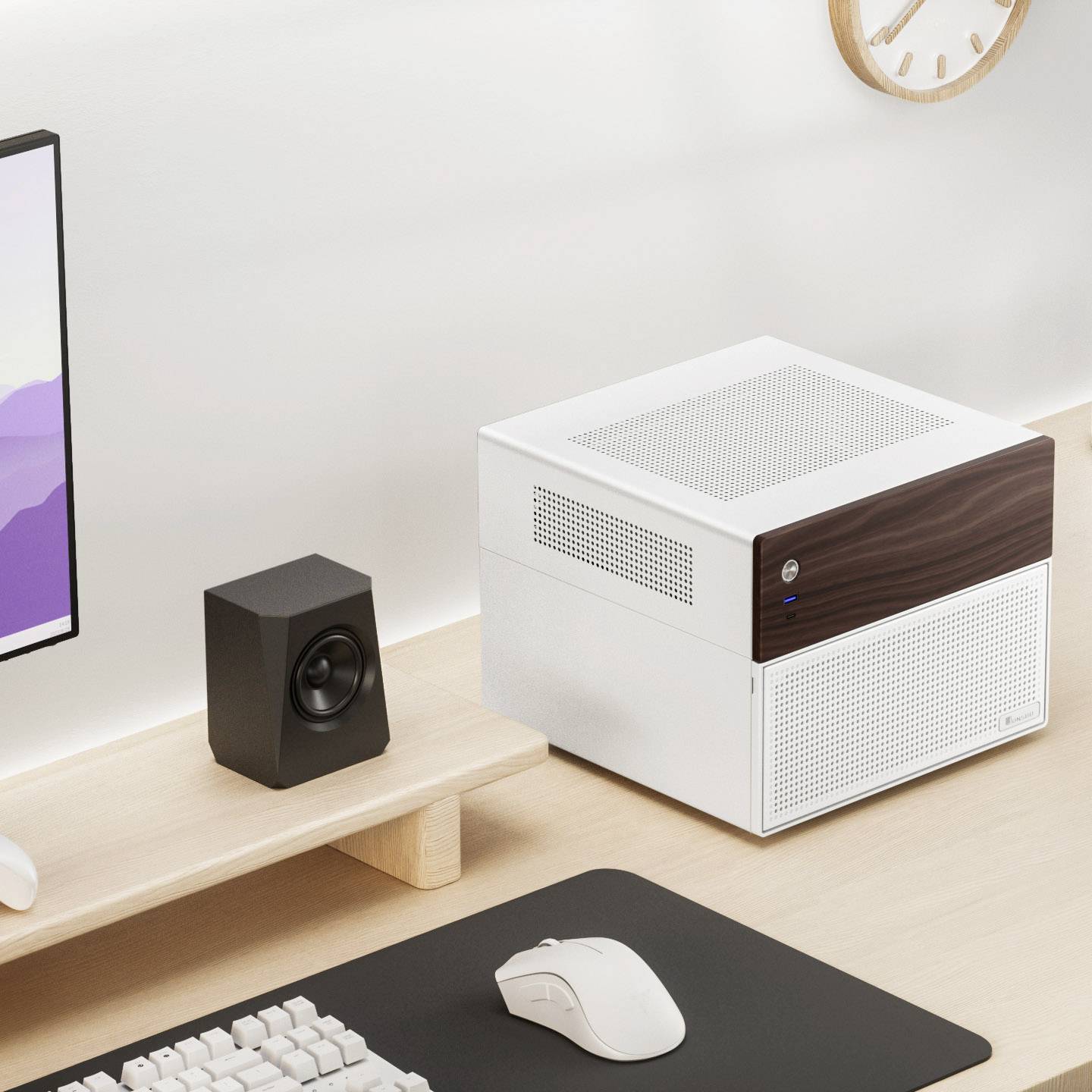 Ein Schreibtisch mit einem kleinen weißen Computer, daneben ein Lautsprecher, eine Maus und eine Tastatur. Wanduhr im Hintergrund.