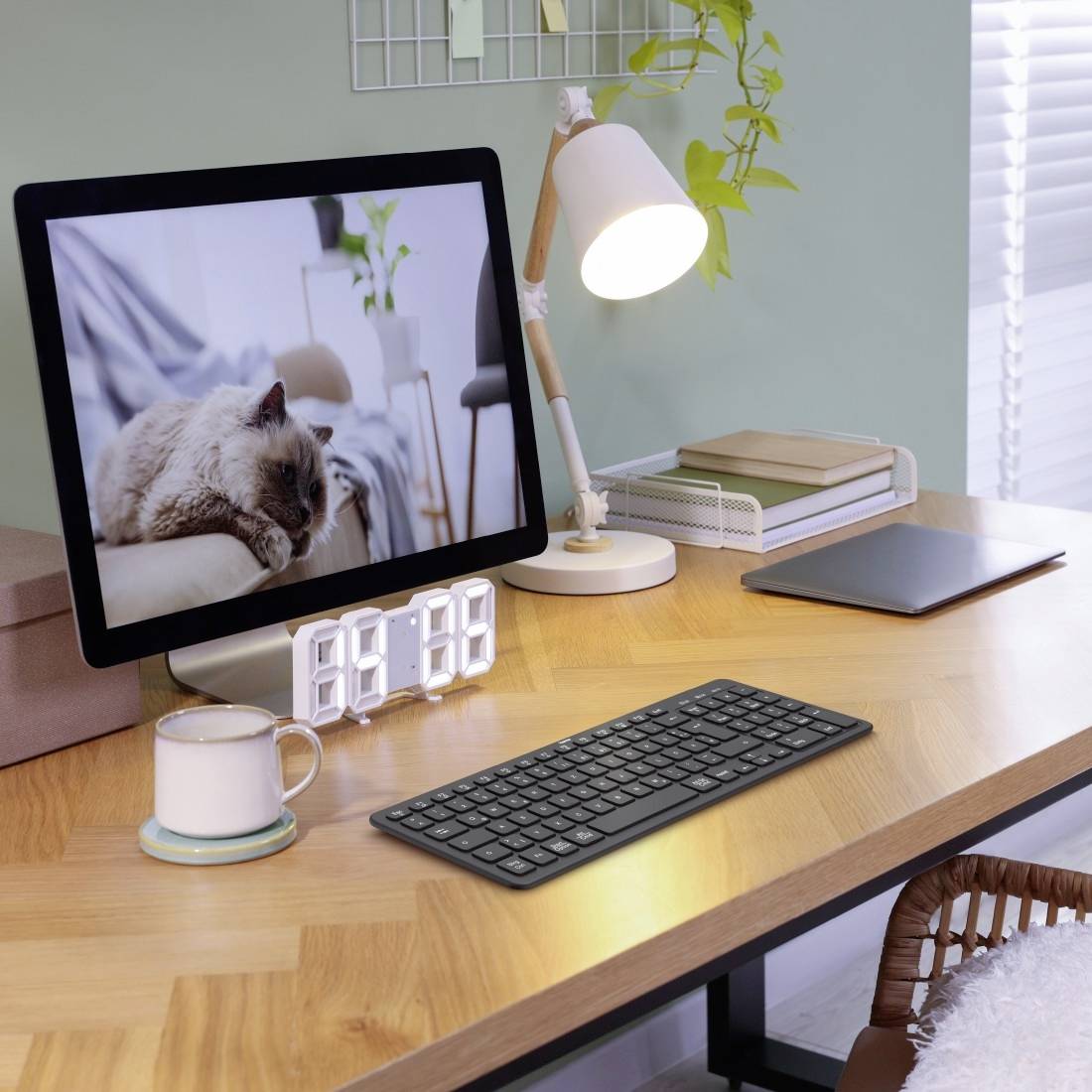 Ein Schreibtisch mit einem Monitor, der eine Katze zeigt. Daneben stehen eine Tischlampe, eine Kaffeetasse, eine digitale Uhr und ein Laptop.