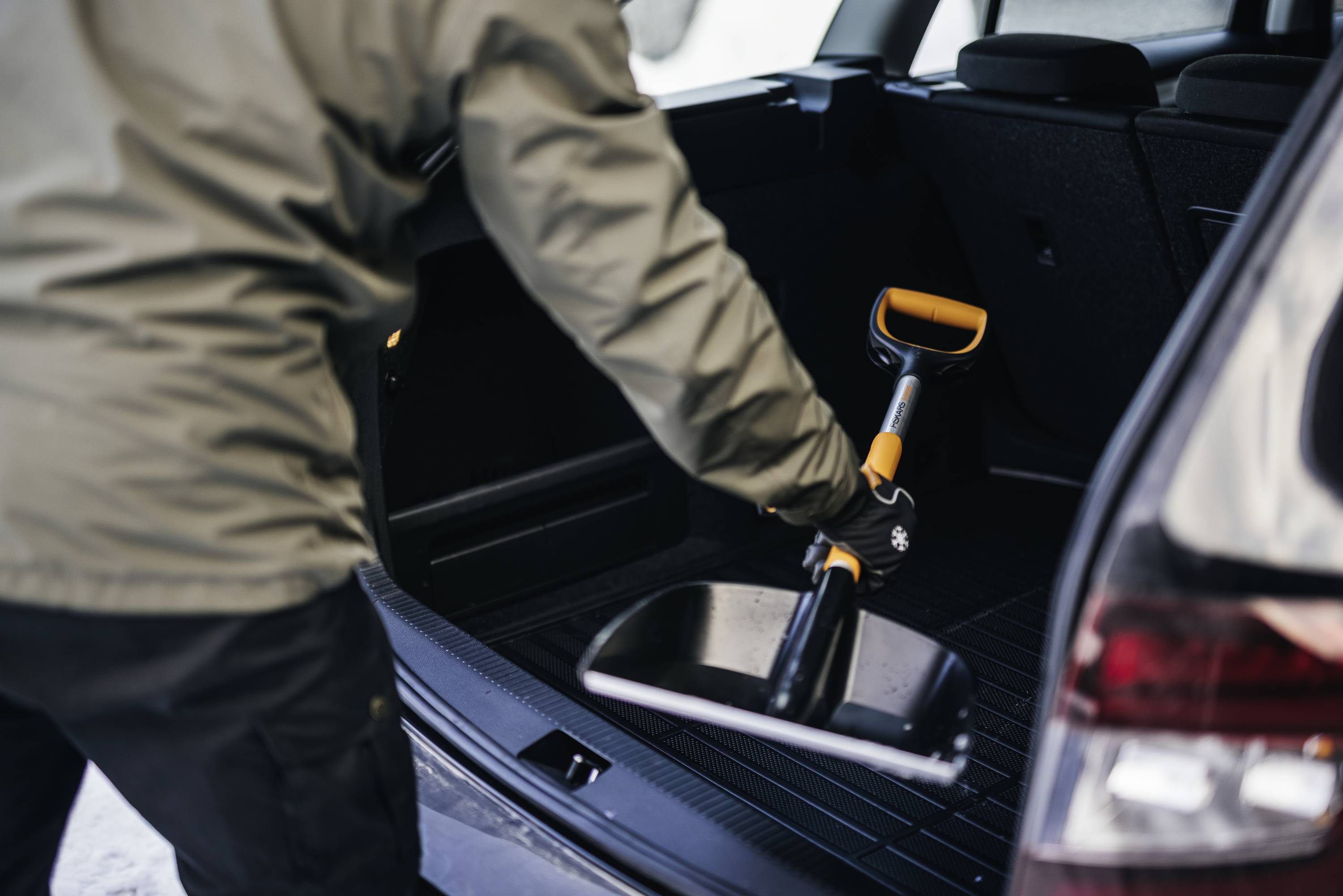 Eine Person legt eine Schaufel in den Kofferraum eines Autos. Die Szene wirkt winterlich, möglicherweise nach Räumarbeiten im Schnee.