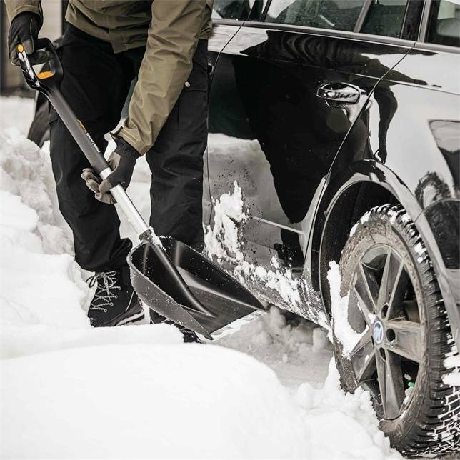 Eine Person schaufelt Schnee neben einem schwarzen Auto. Der Boden ist mit Schnee bedeckt, und das Fahrzeug hat Schneerückstände an den Reifen.