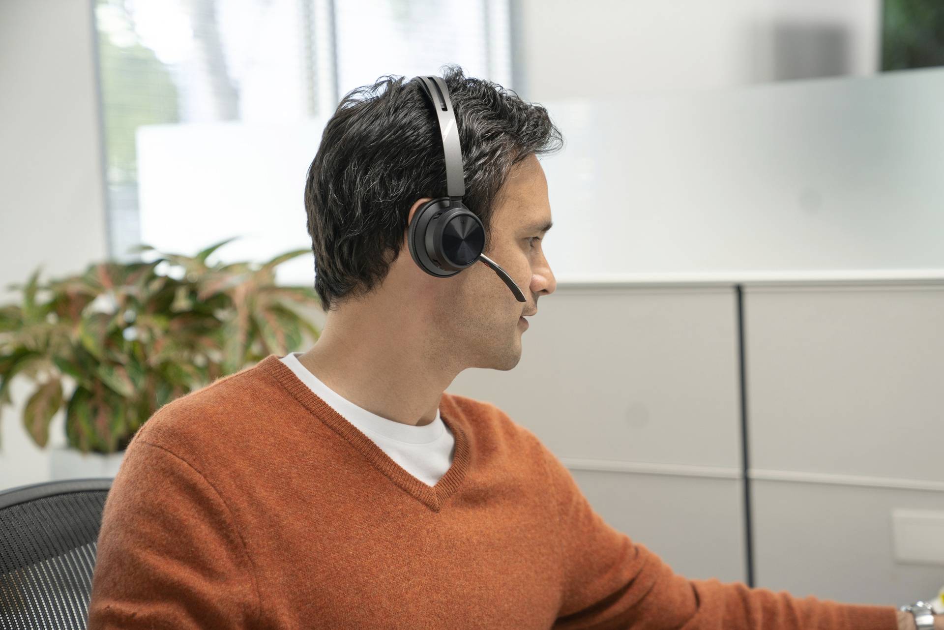 Ein Mann in einem orangefarbenen Pullover sitzt in einem Büro und trägt ein Headset. Im Hintergrund sind Pflanzen zu sehen.