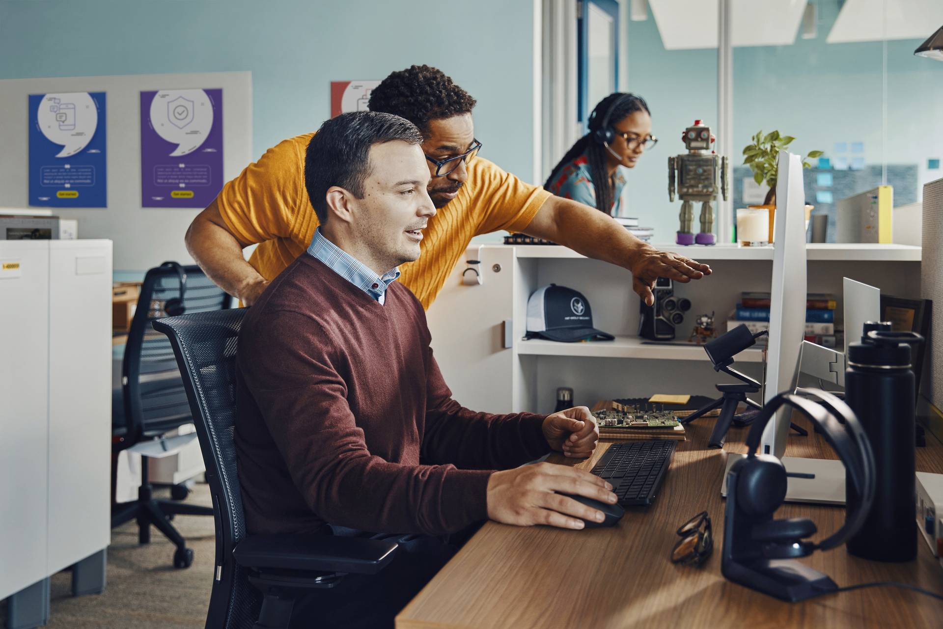 Zwei Personen arbeiten zusammen an einem Computer im Büro. Eine dritte Person im Hintergrund telefoniert und arbeitet an einem Schreibtisch.
