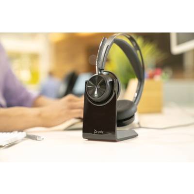 Ein Headset auf einem Tisch mit Ladestation. Im Hintergrund tippt eine Person auf einer Tastatur. Büroumgebung mit Pflanzen.