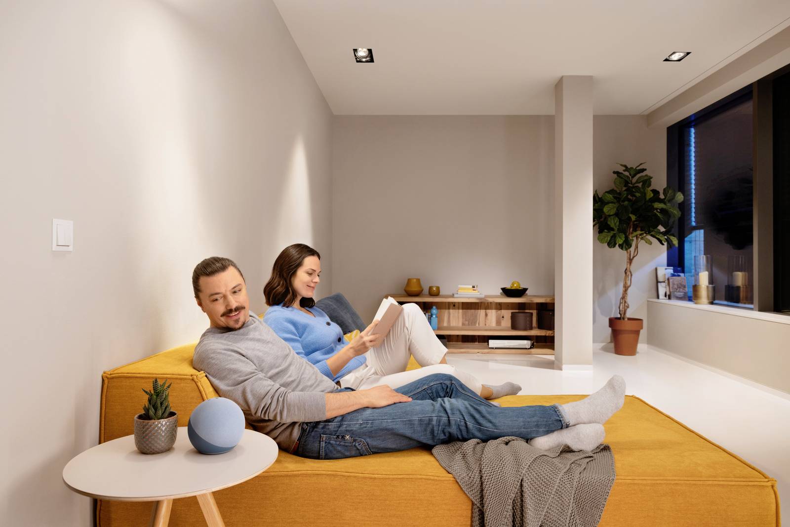 Ein Mann und eine Frau sitzen entspannt auf einem gelben Sofa in einem modernen Wohnzimmer. Die Frau liest ein Buch, eine Pflanze steht im Hintergrund.