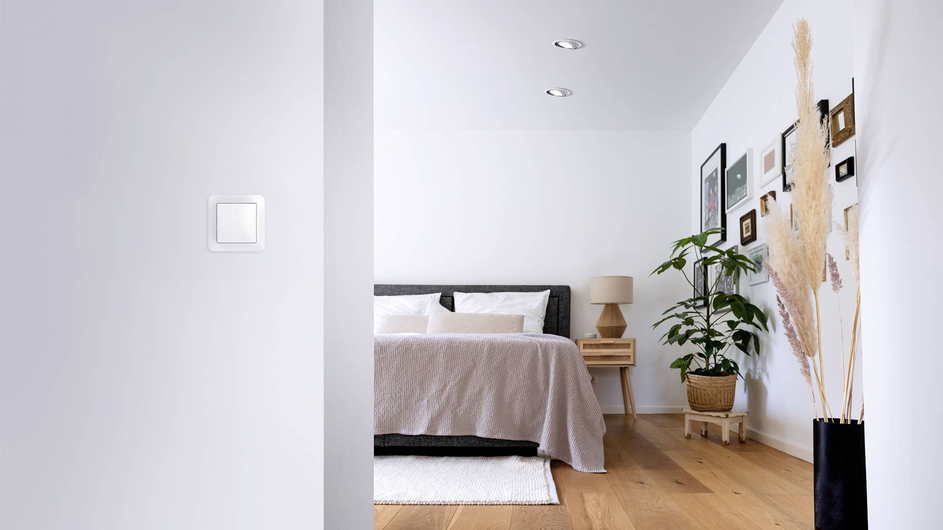 Ein modernes Schlafzimmer mit einem Bett, einer Lampe auf einem kleinen Holztisch, Pflanzen und gerahmten Bildern an der weißen Wand.