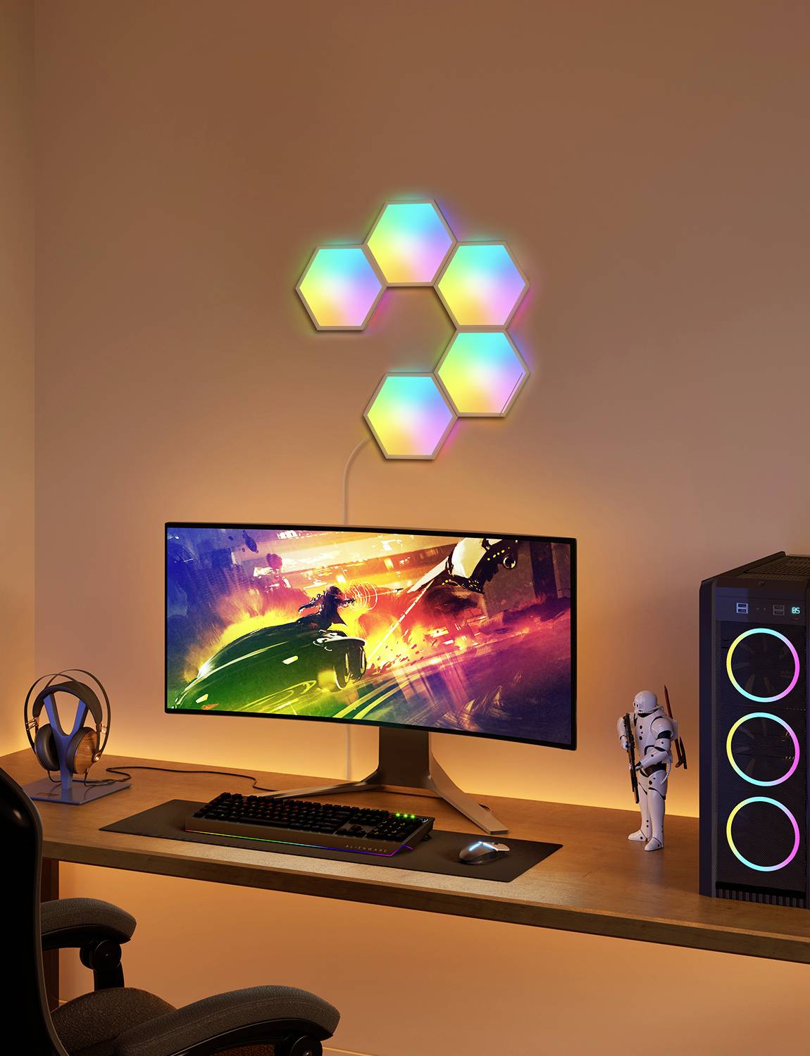Schreibtisch mit beleuchtetem Monitor, bunter Wandleuchte in Wabenform, Kopfhörer, PC-Gehäuse mit leuchtenden Lüftern und Dekoration.