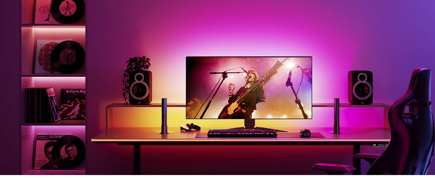Ein moderner Schreibtisch mit einem großen Monitor zeigt ein Konzert. Farbige LED-Leuchten beleuchten die Szene in Pink und Orange. Lautsprecher und Schallplattenregale sind sichtbar.