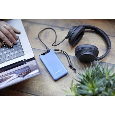 Eine Person arbeitet mit einem Laptop, daneben liegen ein blauer externer Akku und schwarze Kopfhörer auf einem Holztisch. Im Vordergrund ist eine Pflanze.