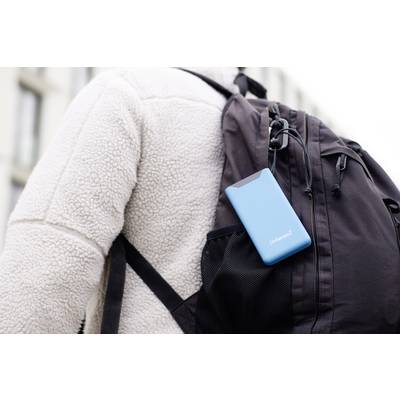 Eine Person mit weißer Jacke trägt einen schwarzen Rucksack, an dem ein blaues tragbares Gerät in einer Außentasche befestigt ist.