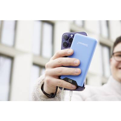 Eine Person hält ein Smartphone mit einer blauen Powerbank, die an das Gerät angeschlossen ist. Hintergrund ist ein Gebäude mit Fenstern.