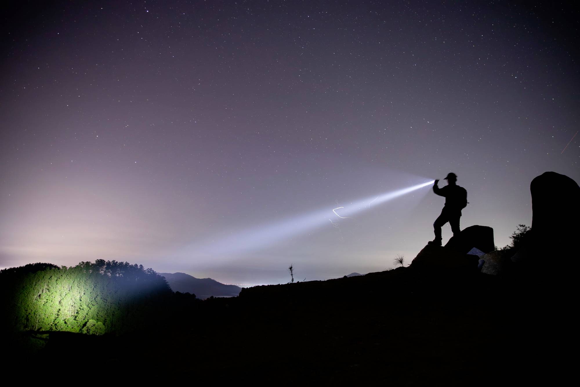 Eine Person leuchtet mit einer Taschenlampe in die Nacht, beleuchtet einen Hügel. Der Himmel ist klar mit sichtbaren Sternen.