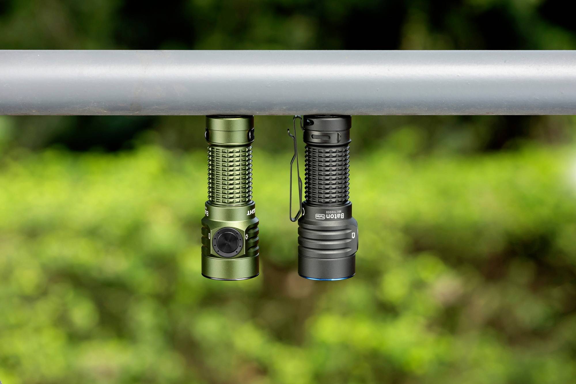 Zwei Taschenlampen hängen nebeneinander an einer Metallstange; eine ist grün, die andere schwarz. Im Hintergrund sind verschwommene Bäume.