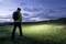 Eine Person mit Rucksack und Stirnlampe wandert bei Dämmerung über eine Wiese Richtung Horizont, im Hintergrund leicht bewölkter Himmel.