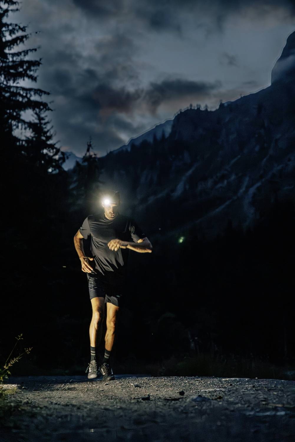 Ein Mann joggt bei Nacht auf einem Waldweg, beleuchtet durch eine Stirnlampe. Berge und Bäume im Hintergrund, dunkler Himmel.