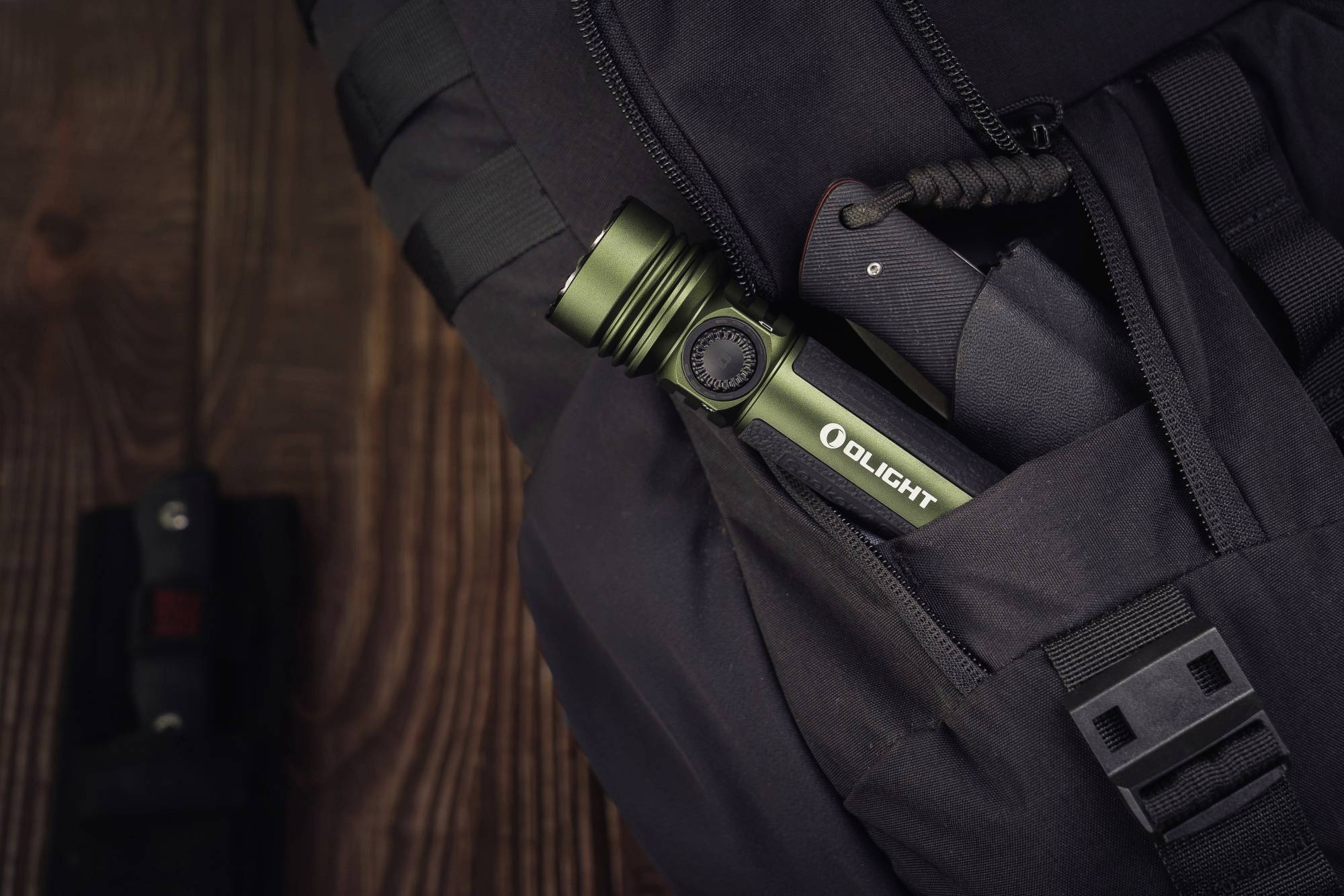 Eine grüne Taschenlampe und ein Messer stecken in einer schwarzen Tasche. Auf Holzuntergrund ist eine weitere Messertasche zu sehen.