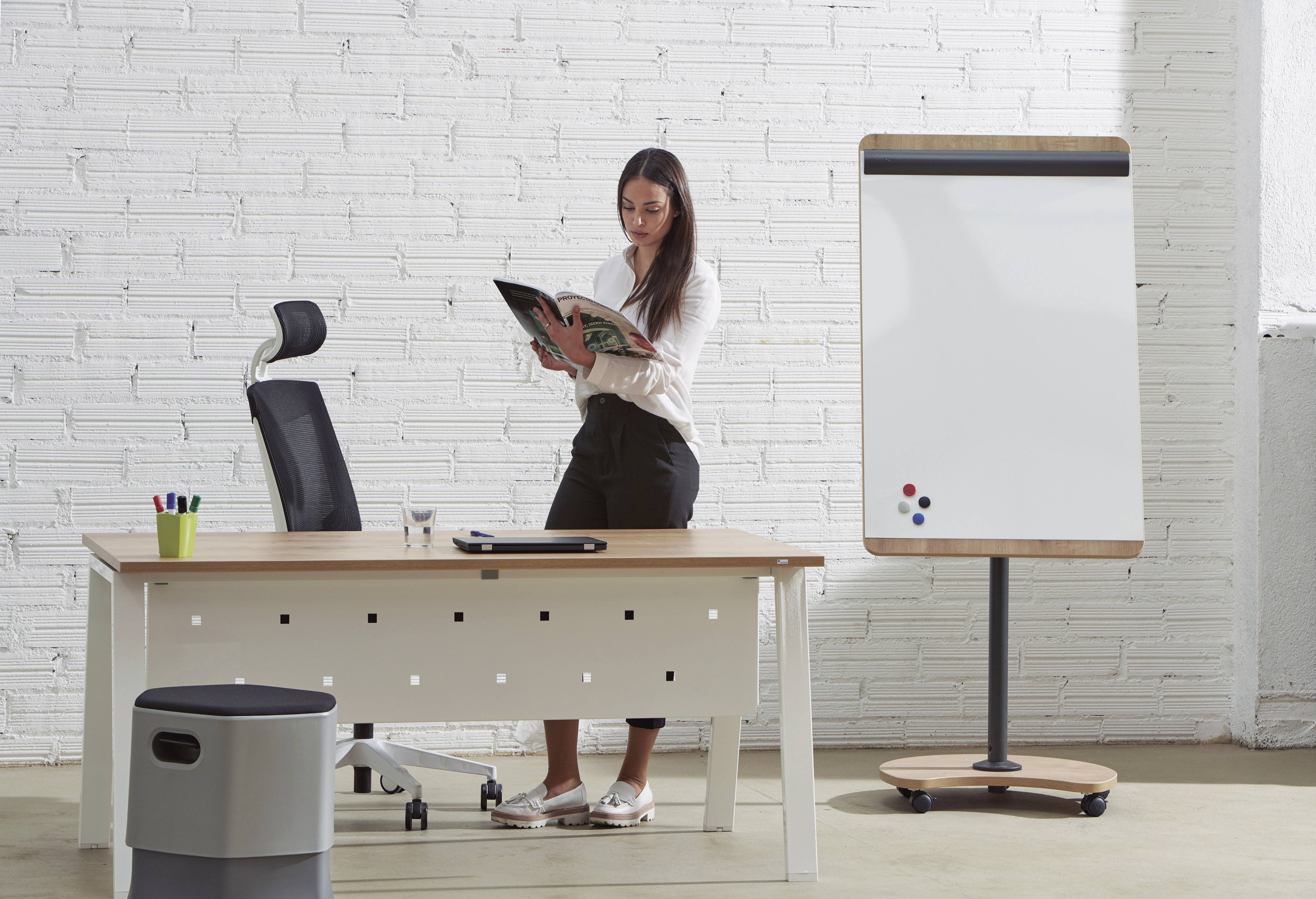 Eine Frau steht in einem modernen Büro und liest ein Magazin neben einem Schreibtisch und einem leeren Flipchart.