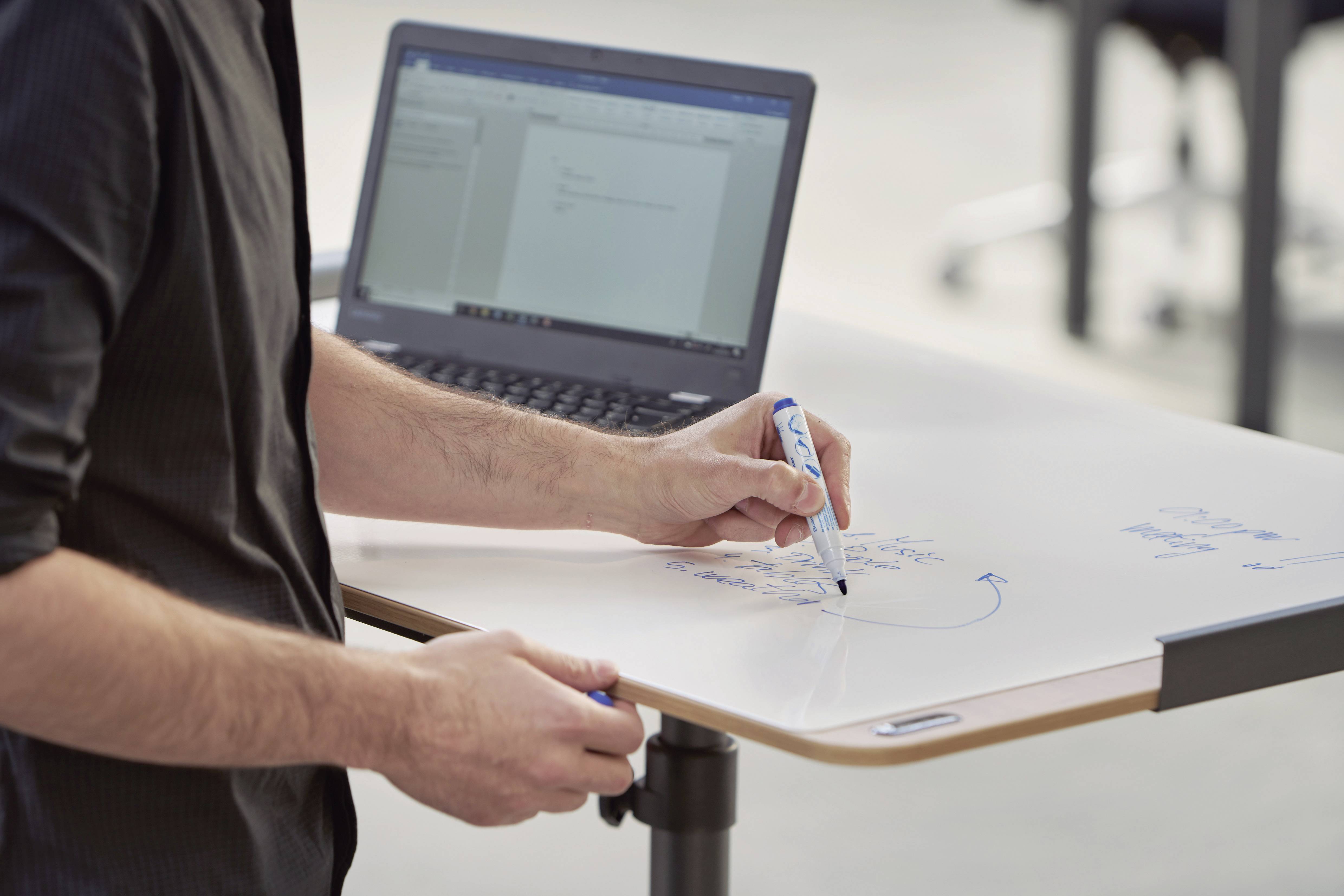 Eine Person schreibt mit einem blauen Stift auf einem höhenverstellbaren Tisch. Im Hintergrund ist ein aufgeklappter Laptop zu sehen.