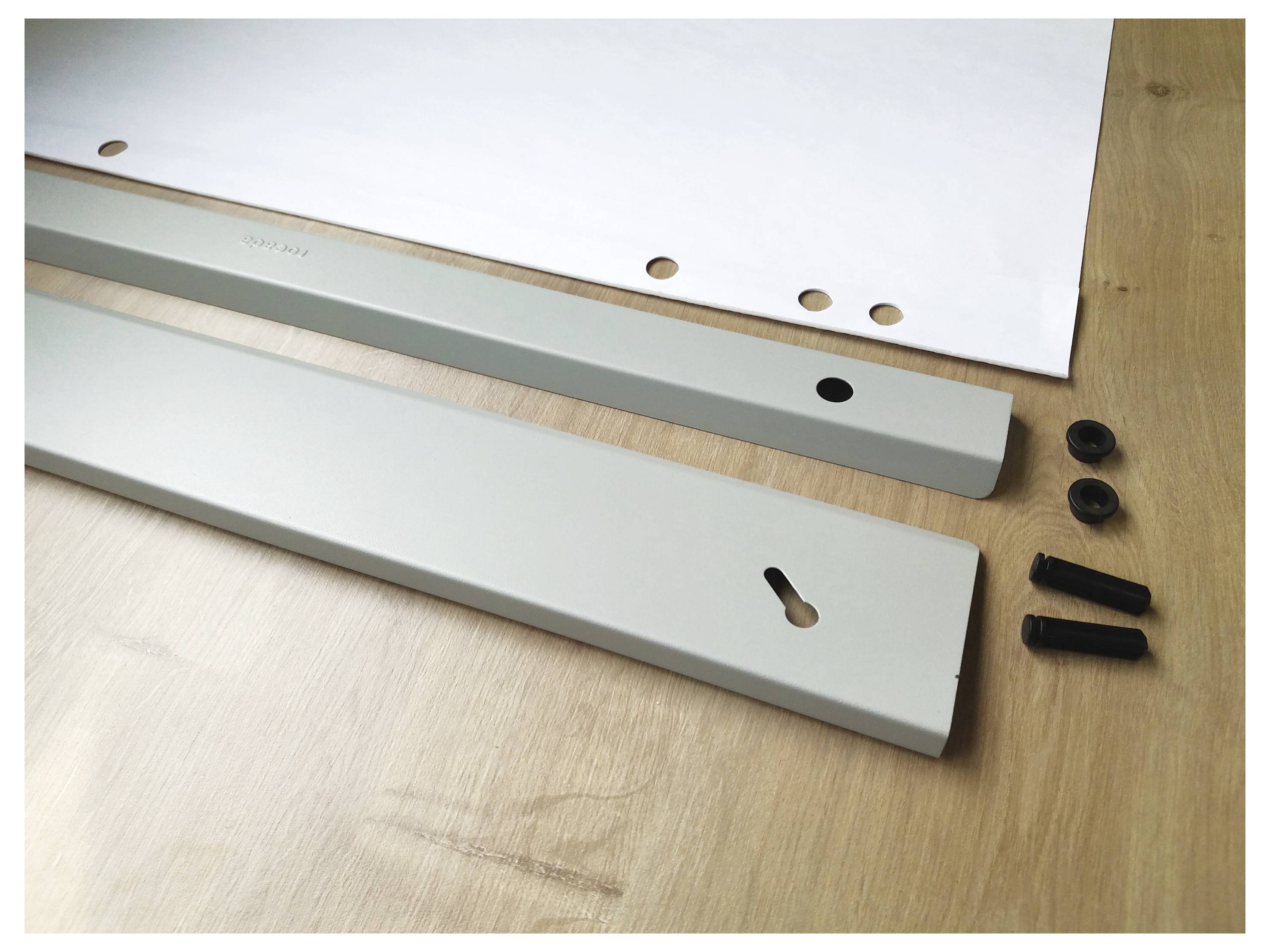 Weiße Metallstreben und Schrauben liegen auf einem Holztisch neben einem Papier mit Lochungen, möglicherweise Teile zur Montage.