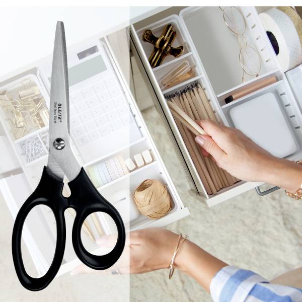 Eine Person arrangiert Schreibtischzubehör in einer Schublade, darunter Büroklammern, Stifte und Notizblöcke. Im Vordergrund liegt eine Schere.