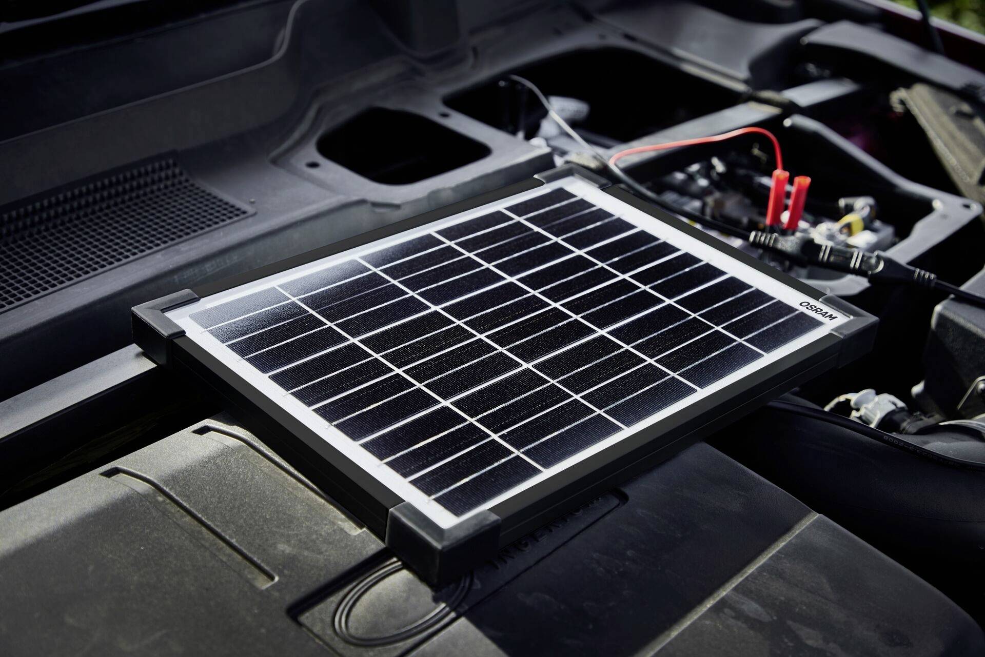 Ein Solarpanel liegt auf dem offenen Motorraum eines Autos und ist mit Kabeln angeschlossen, wahrscheinlich zur Energieversorgung der Batterie.