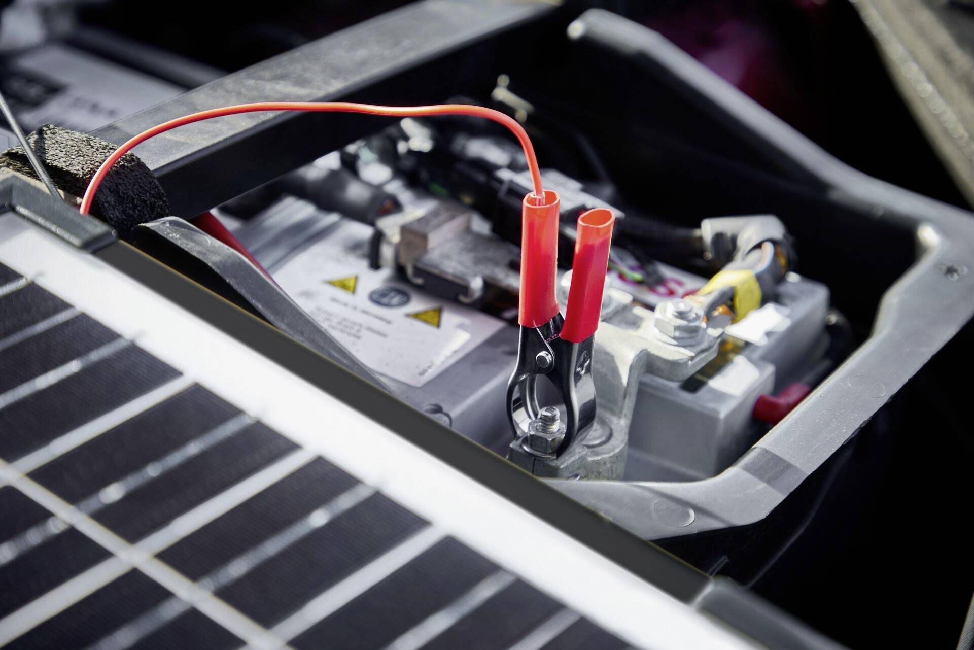 Autobatterie mit angeschlossenem roten Überbrückungskabel, das an einem Solarpanel angebracht ist. Szenario deutet auf Laden oder Starthilfe.